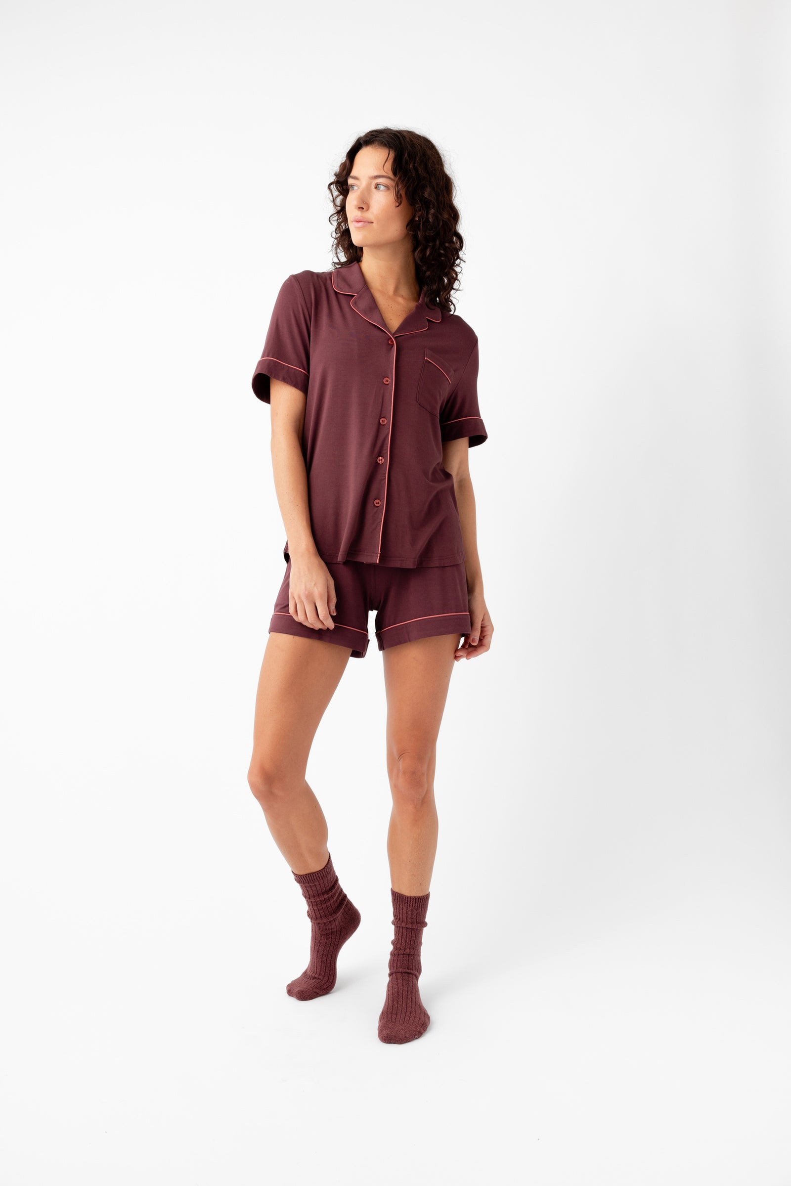 A woman with curly hair stands against a white background wearing Cozy Earth's Women's Bamboo Stretch-Knit Short Sleeve Pajama Set in burgundy with matching socks, looking to the side with a relaxed expression.