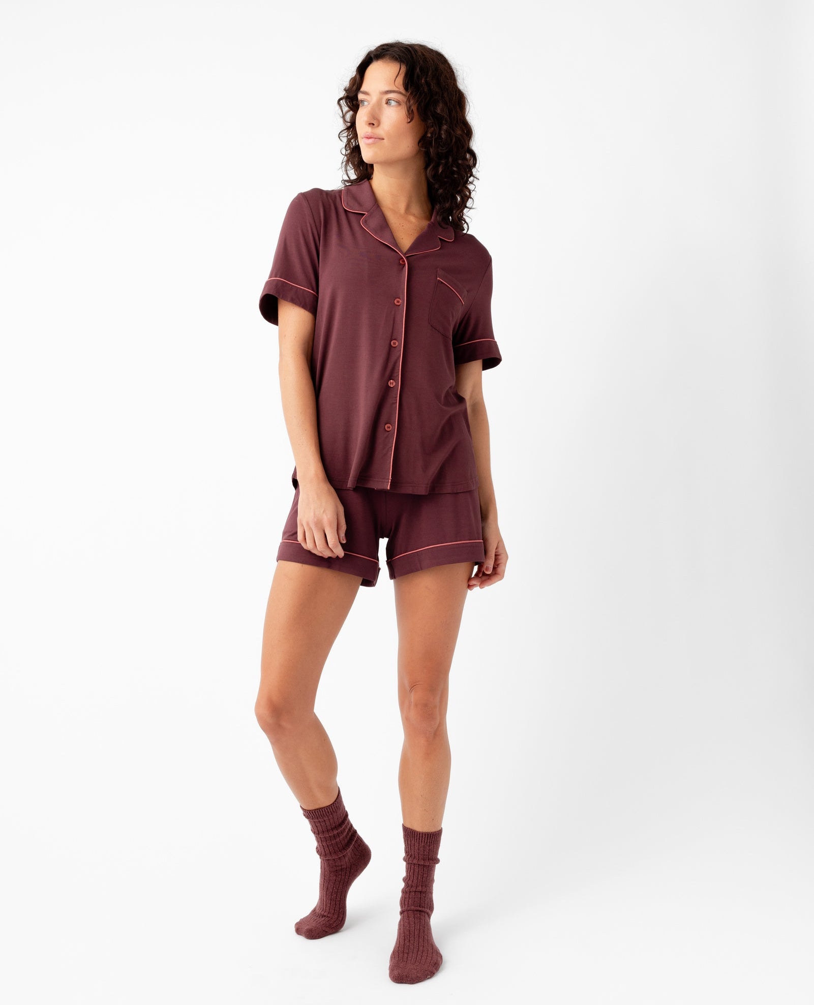 A woman with curly hair stands against a white background wearing Cozy Earth's Women's Bamboo Stretch-Knit Short Sleeve Pajama Set in burgundy with matching socks, looking to the side with a relaxed expression.|Color:Burgundy