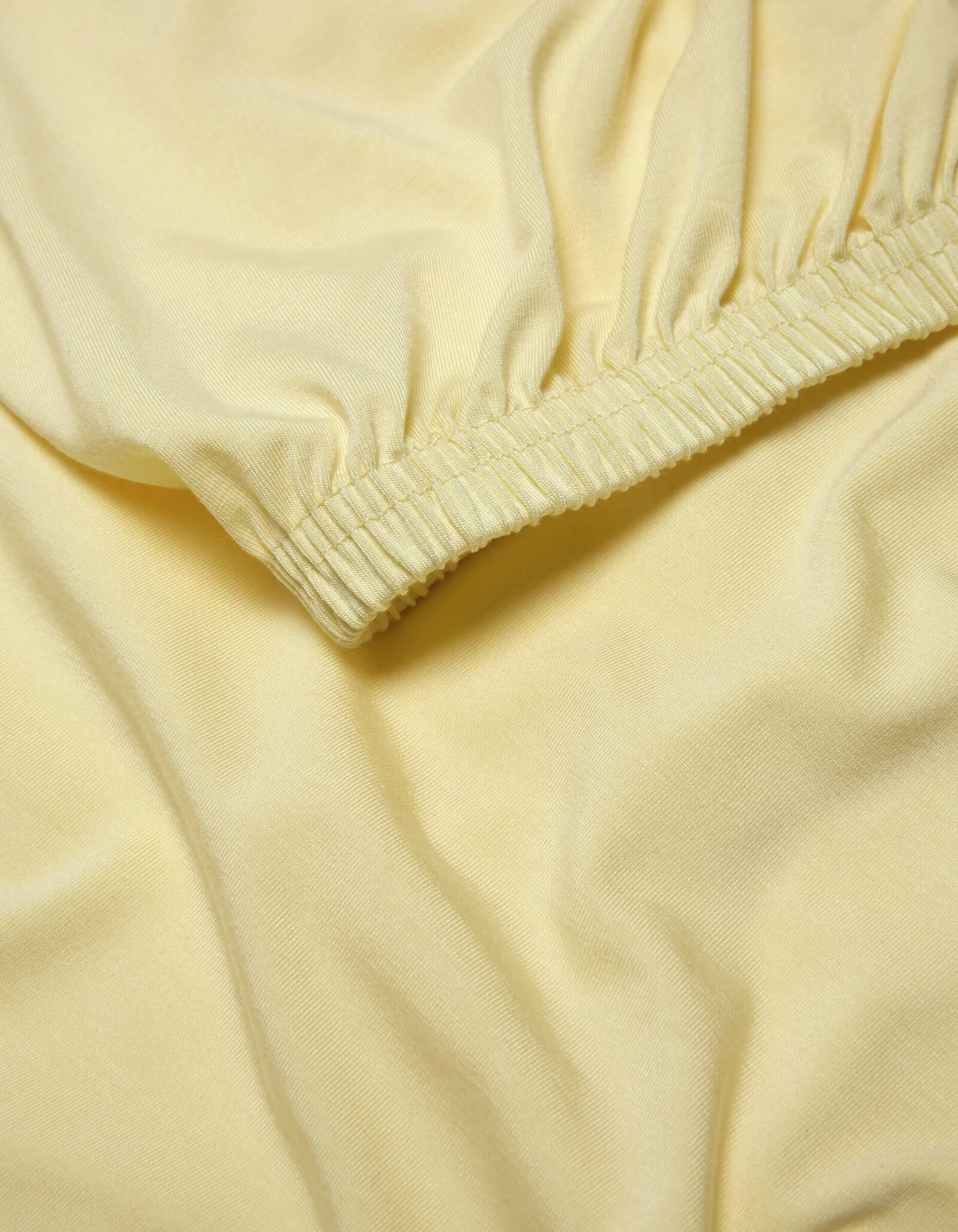 A close-up of the Cozy Earth Bamboo Jersey Fitted Sheet shows light yellow fabric with elasticized, gathered edges that create gentle folds and a smooth, soft texture.