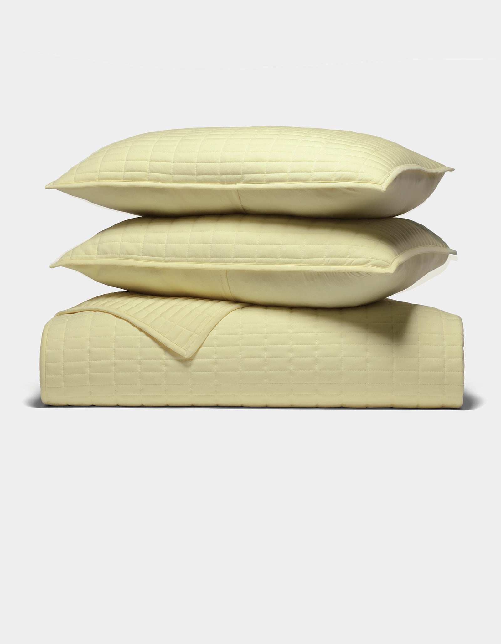 The Cozy Earth Bamboo Jersey Quilt Bundle features two pale yellow pillows stacked on a matching quilted blanket, all with a subtle square pattern, neatly arranged against a plain white background.