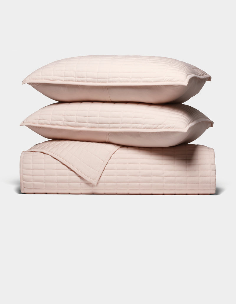 The Cozy Earth Bamboo Jersey Quilt Bundle features two light pink quilted pillows stacked atop a matching quilted blanket, all neatly arranged against a plain white background.