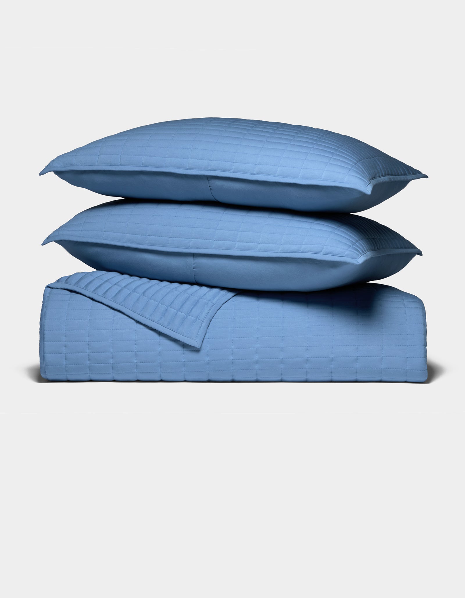 Two blue pillows rest atop a matching blue Bamboo Jersey Quilt Bundle by Cozy Earth, all showcasing a square grid pattern, set against a plain white background.