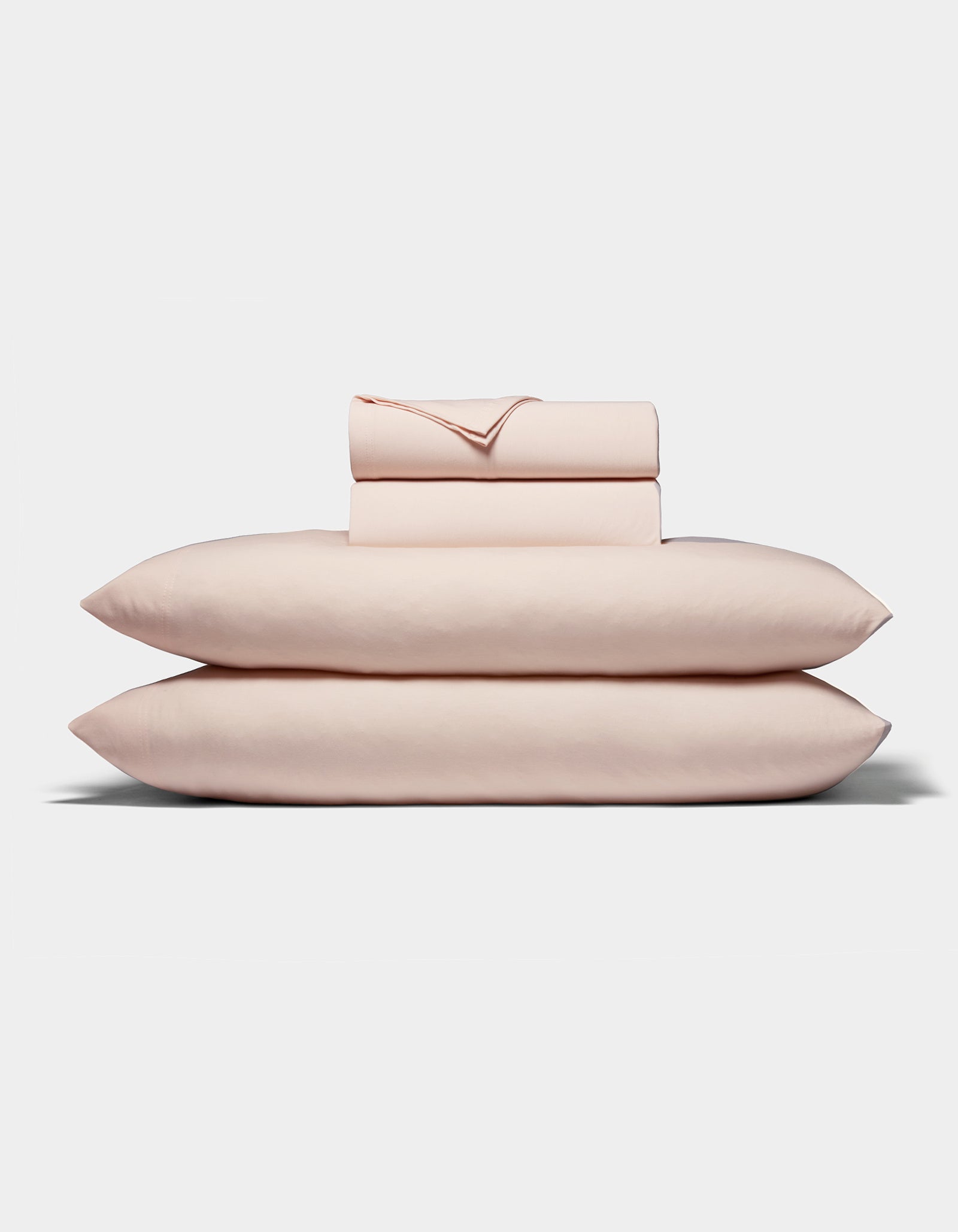 A neatly folded stack of Cozy Earth's Bamboo Jersey Sheet Set, in pale pink, featuring two pillows, a fitted sheet, a flat sheet, and pillowcases arranged on a white background.