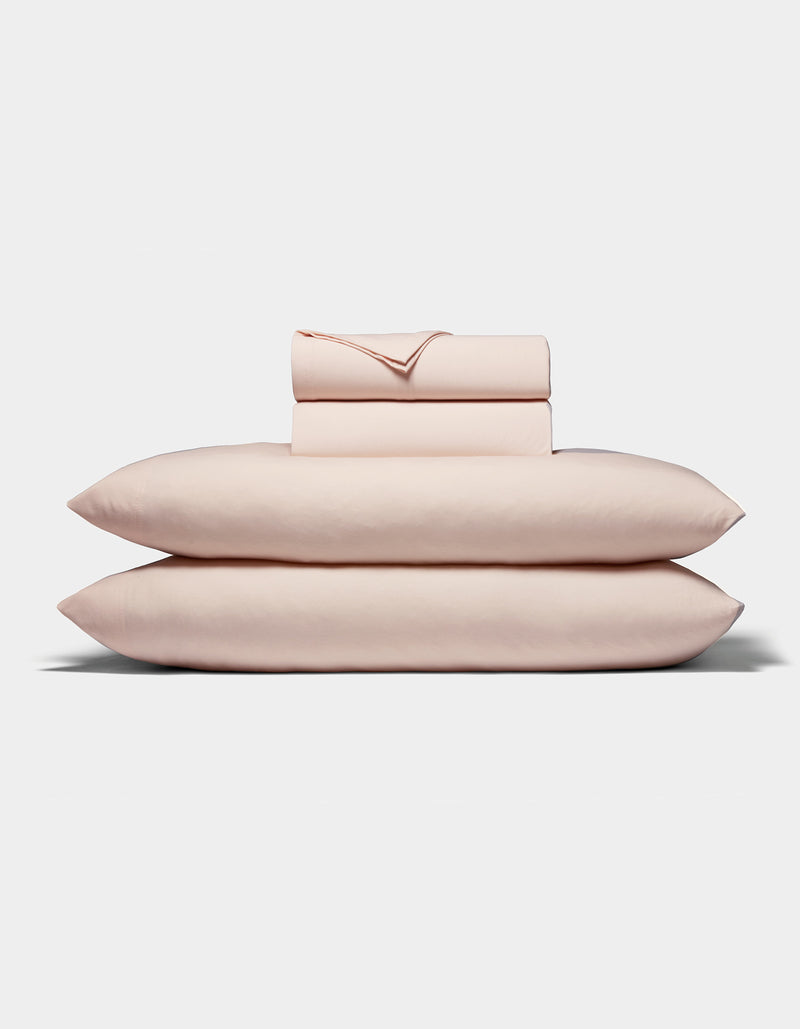 A neatly folded stack of Cozy Earth's Bamboo Jersey Sheet Set, in pale pink, featuring two pillows, a fitted sheet, a flat sheet, and pillowcases arranged on a white background.