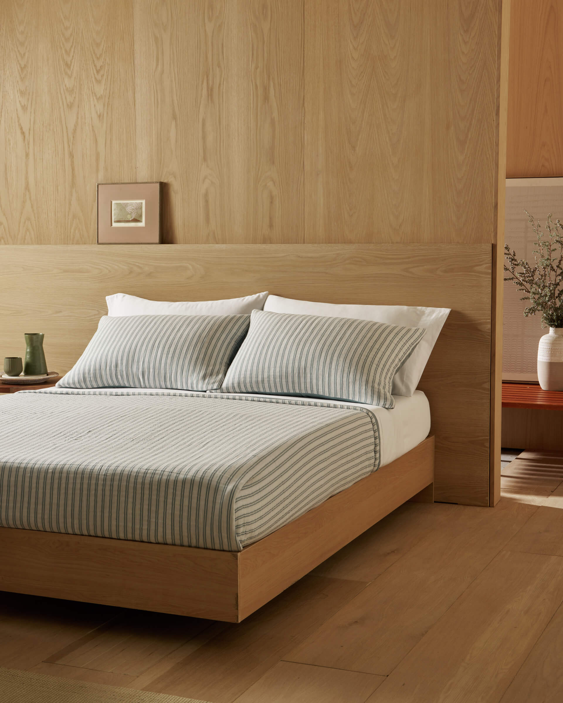 A minimal bedroom with light wood walls and floor, featuring a wooden bed styled with Cozy Earth’s Waterfront Coverlet, two pillows, a side table holding cups, and a vase with branches in the corner.