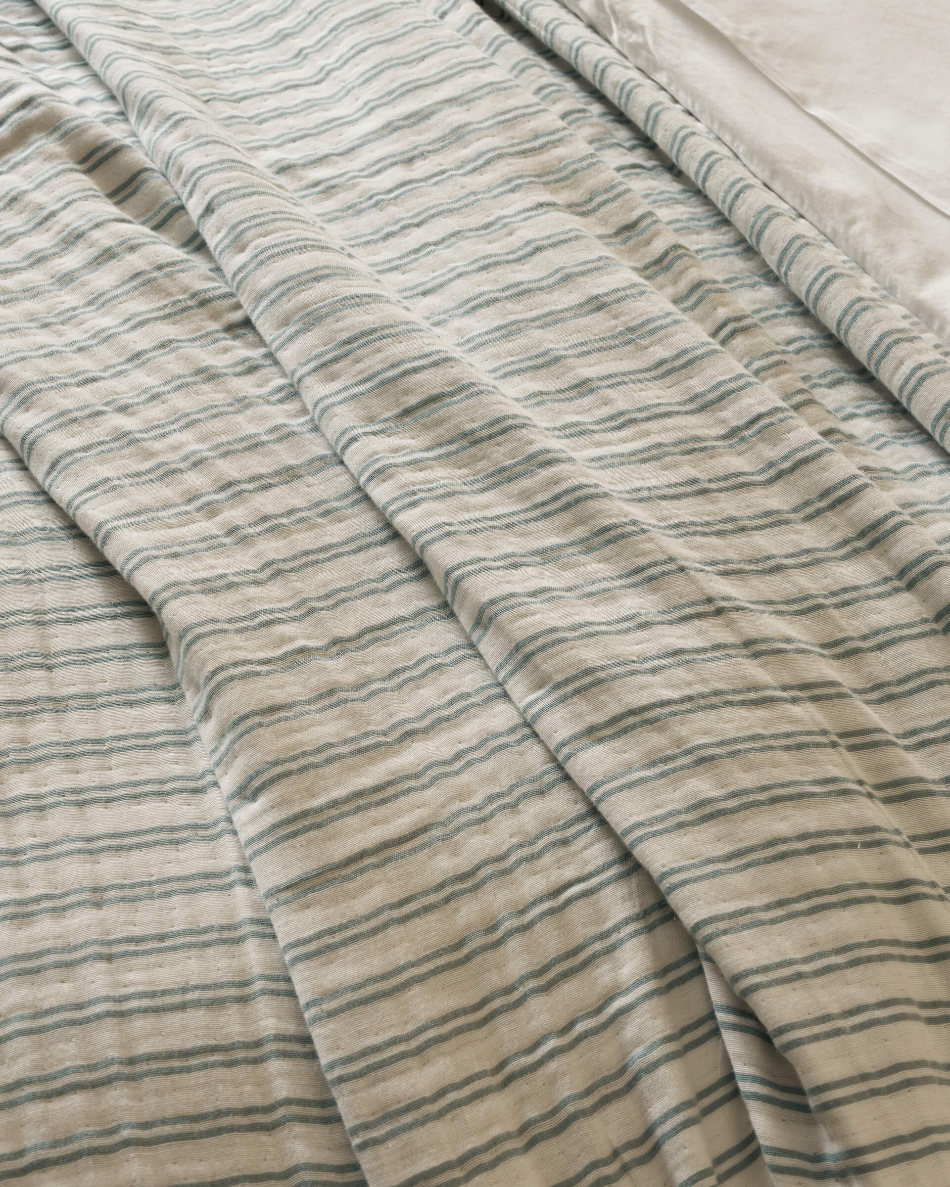 The Waterfront Coverlet by Cozy Earth is shown on a bed, displaying its cream color with thin blue-green stripes and gentle folds. A smooth white sheet is visible at the top right corner.