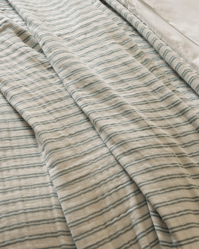 The Waterfront Coverlet by Cozy Earth is shown on a bed, displaying its cream color with thin blue-green stripes and gentle folds. A smooth white sheet is visible at the top right corner.