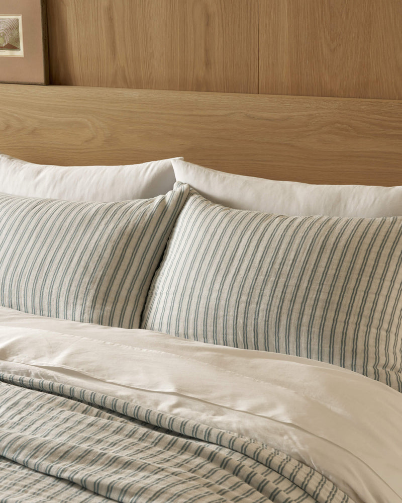 A bed made with Cozy Earth Waterfront Shams features white pillows and blue-and-white striped pillowcases, set against a wooden headboard with a framed picture partially visible on the wall.