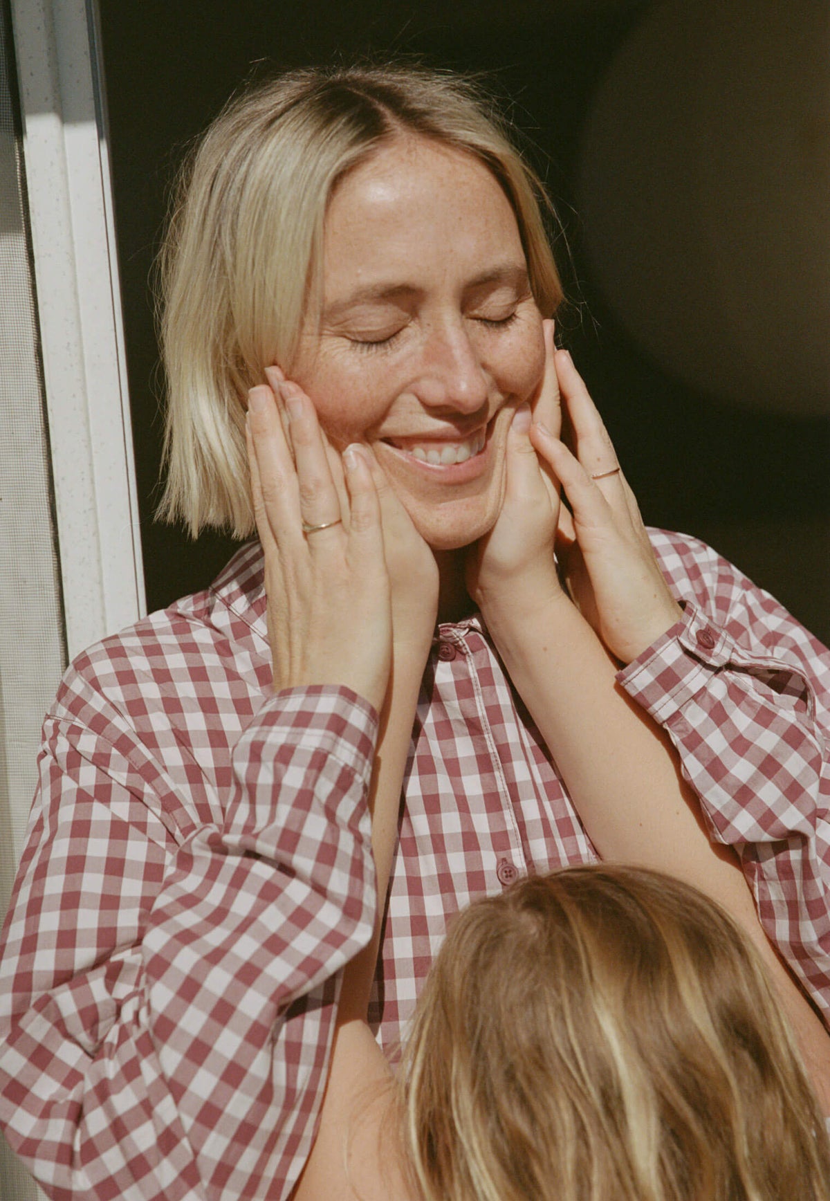 Woman in a red and white checkered shirt holding her face with both hands, smiling.