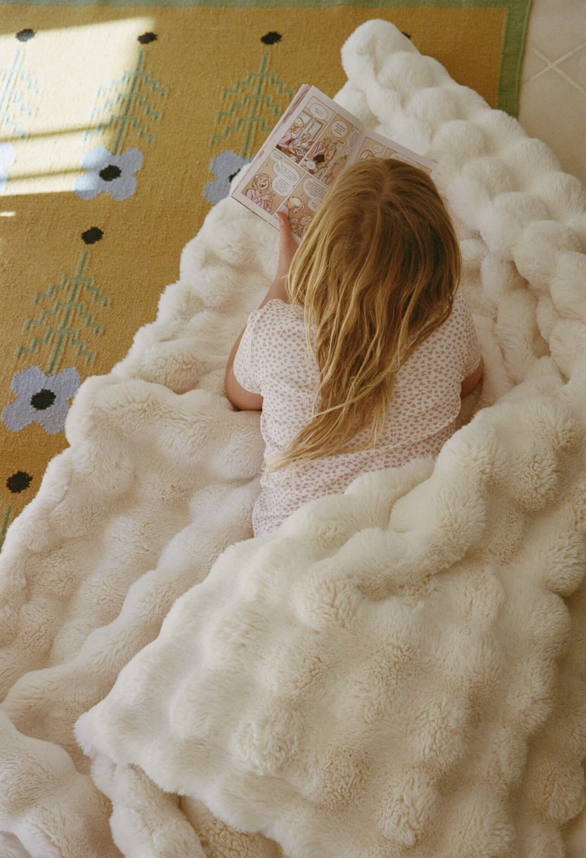 A little girl lying on a fluffy white blanket reading a book.