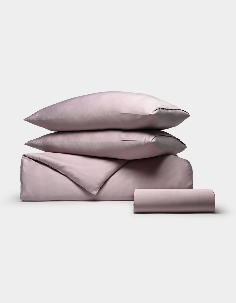 The Cozy Earth Bamboo Bedding Essential Bundle is displayed on a plain white background, featuring a neatly stacked set of light mauve bedding that includes two pillows, a folded sheet, and a rolled sheet.|Color:Lavender