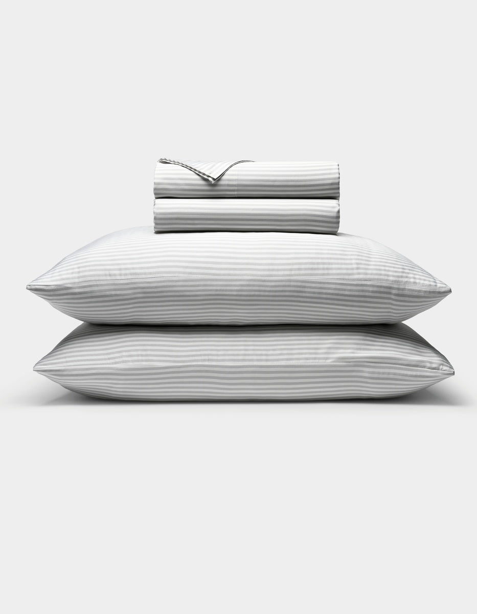 Two stacked pillows with subtle gray and white stripes are shown beneath a neatly folded Cozy Earth Bamboo Sheet Set, all displayed against a plain white background.