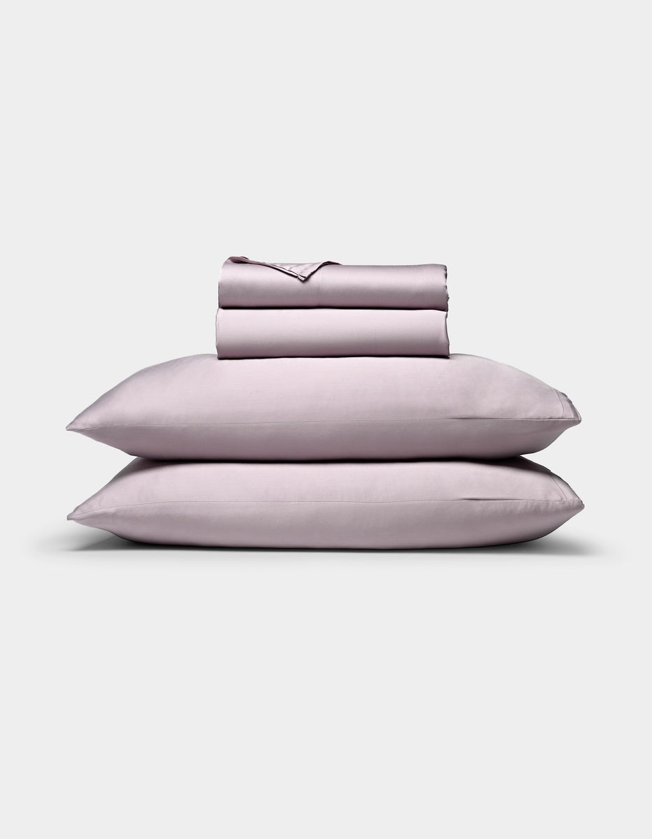 A neatly folded Bamboo Sheet Set by Cozy Earth in light lavender, featuring two stacked pillows, a fitted sheet, a flat sheet, and pillowcases, displayed against a plain white background.
