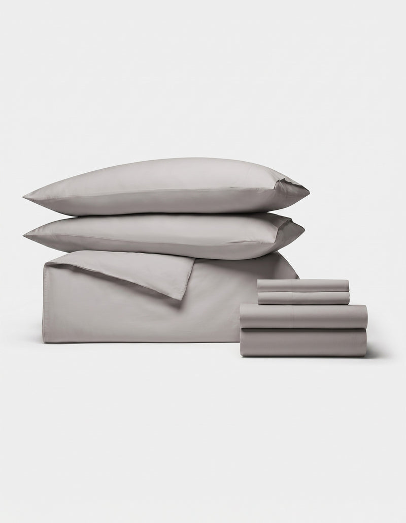 The Cozy Earth Soft-Wash Cotton Bedding Core Bundle features two pillows, a folded duvet, and two sets of folded sheets in light gray, all neatly stacked and displayed on a plain white background.