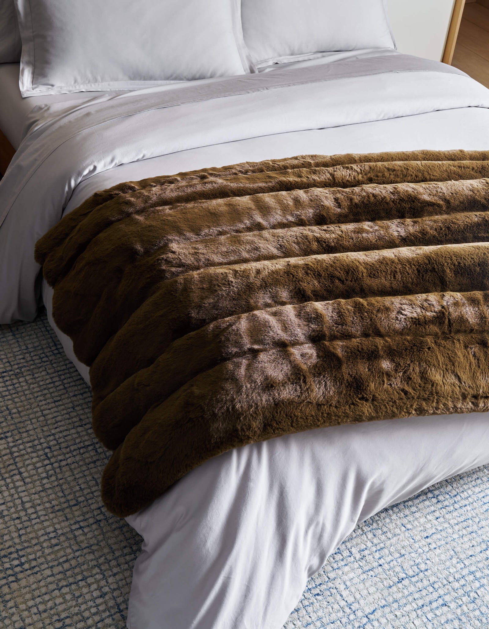 A neatly made bed with white sheets and pillows features the plush Cozy Earth Channel Cuddle Blanket in brown draped across the foot, on top of a blue and white textured rug.