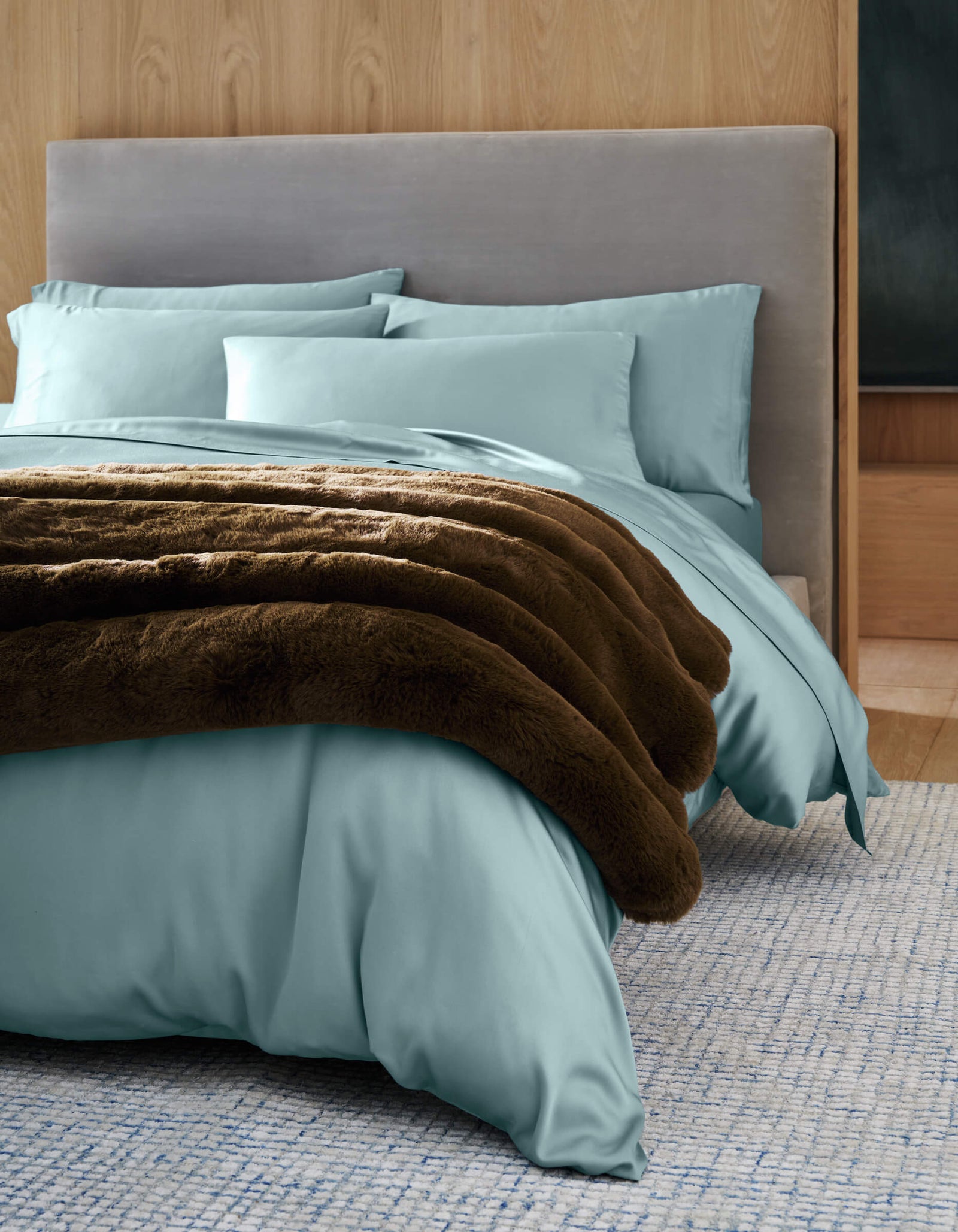 A neatly made bed features a Cozy Earth Channel Cuddle Blanket, light blue bedding, matching pillows, and a soft gray upholstered headboard against a light wood paneled wall on a textured blue rug.