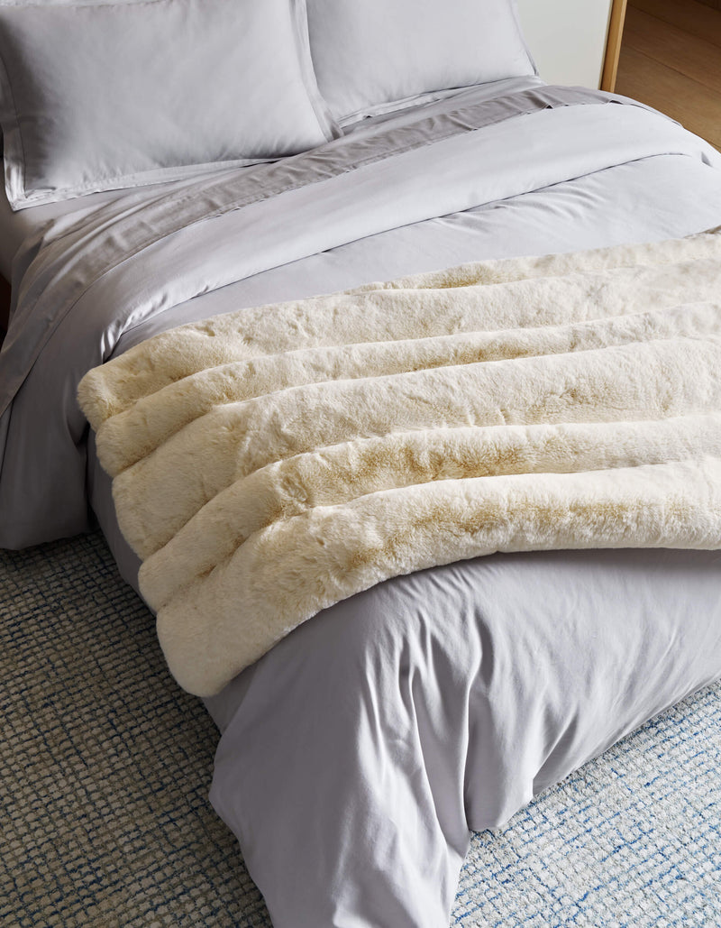 A neatly made bed with light gray sheets and pillowcases is topped with Cozy Earth’s Channel Cuddle Blanket in cream, draped at the foot. A blue and white textured rug is partially visible on the floor.