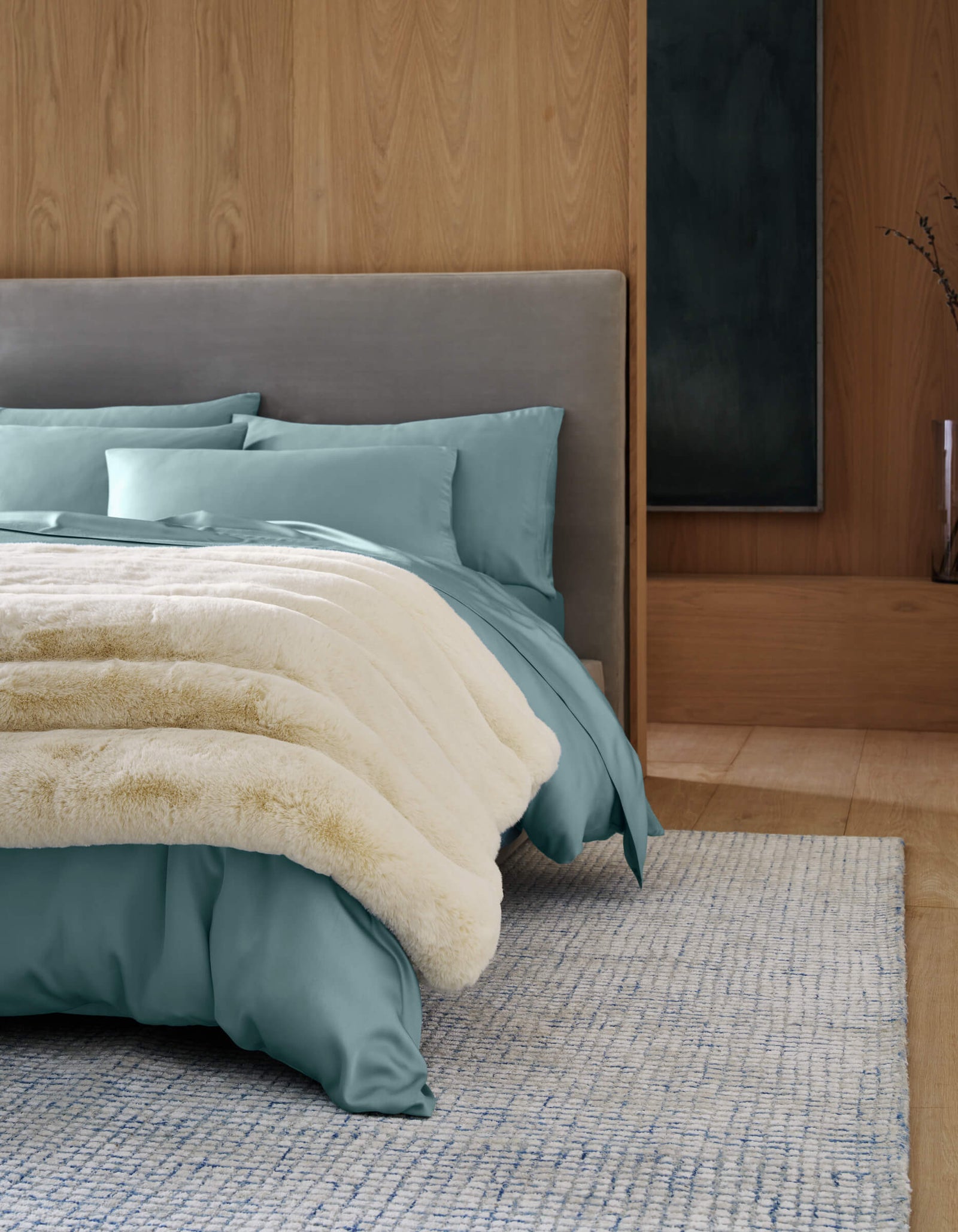 A modern bedroom with a light gray upholstered bed, teal bedding, and the Cozy Earth Channel Cuddle Blanket in cream. Wooden walls, a light blue textured rug, minimal decor, and a tall vase with branches complete the look.