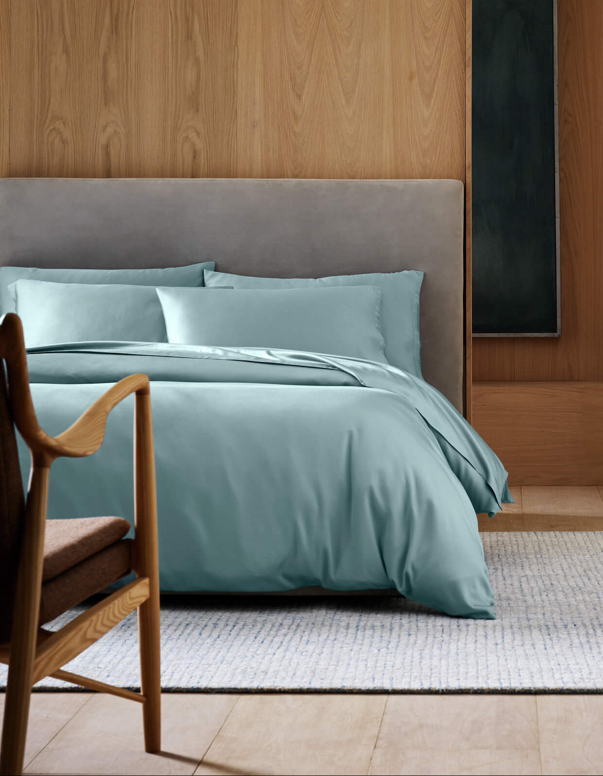 A modern bedroom featuring Cozy Earth's Bamboo Duvet Cover in light blue, a gray upholstered headboard, light wooden wall, wooden chair with brown cushion, and a light woven rug on a wooden floor.