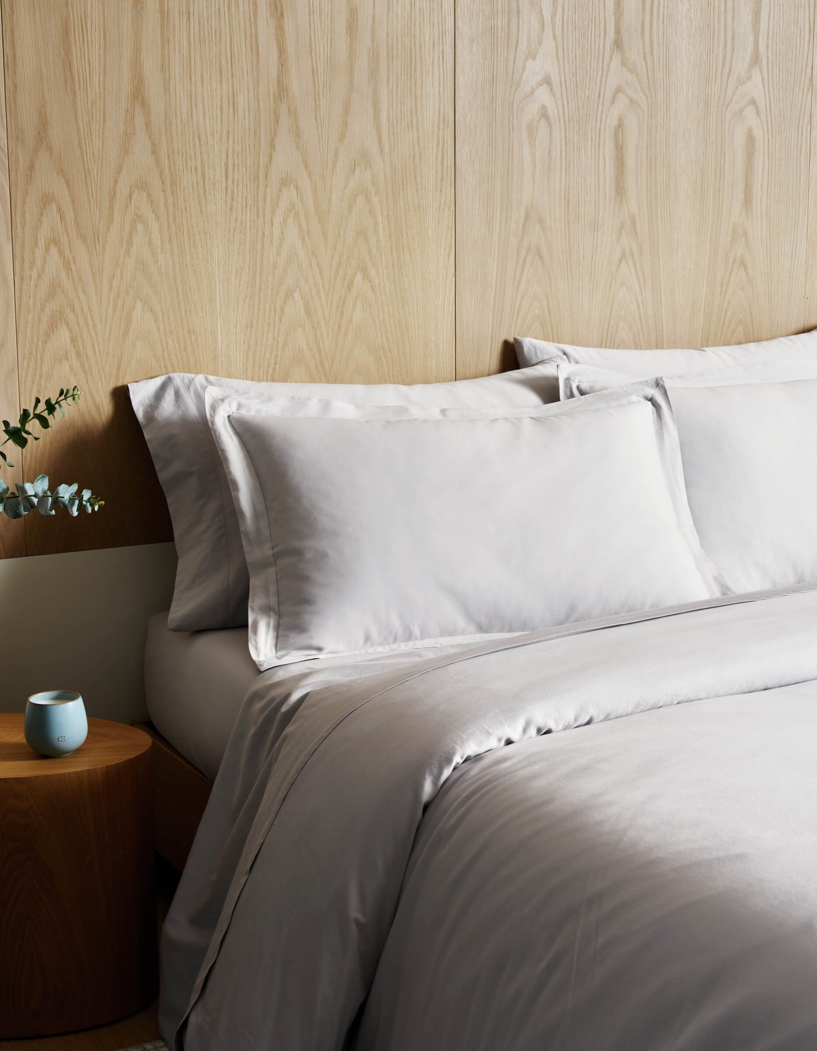 A bed featuring the Cozy Earth Soft-Wash Cotton Duvet Cover in light gray is set against a wood-paneled wall. A small round wooden nightstand displays a blue candle and greenery arranged in a white vase.