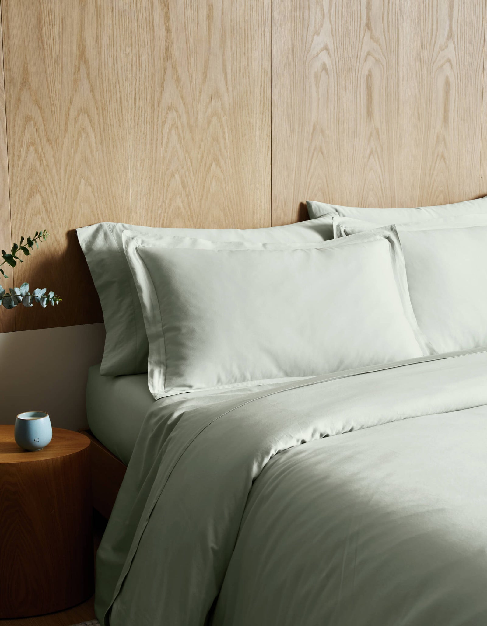 A bed dressed in Cozy Earth's Soft-Wash Cotton Duvet Cover in light green sits beside a round wooden nightstand holding a blue candle and eucalyptus, all set against a light wood-paneled wall.