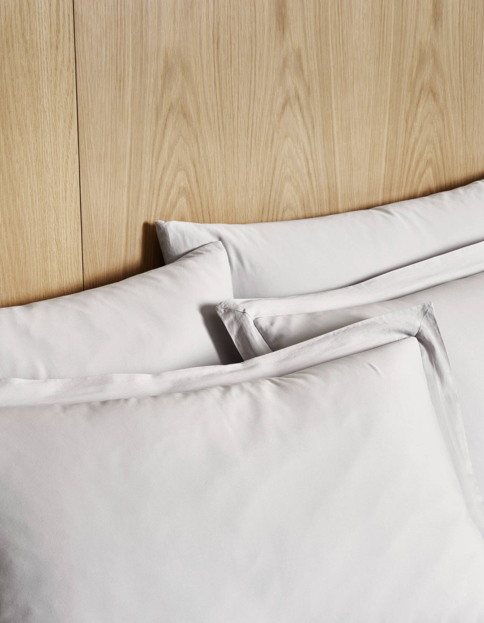 Three Cozy Earth Soft-Wash Cotton Shams in white, featuring smooth fabric, are stacked against a wooden headboard for a clean, minimalistic bed look.