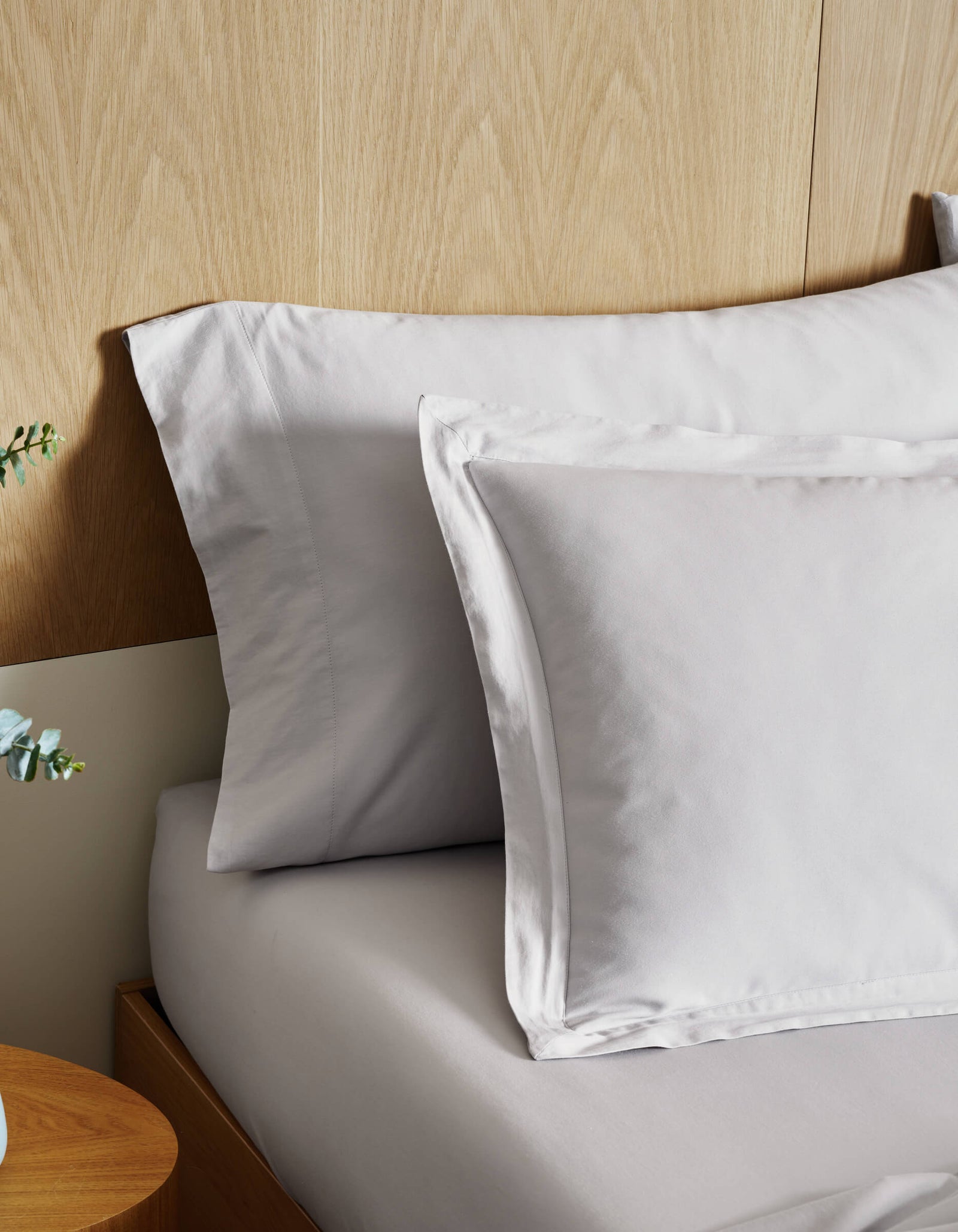 A close-up of a neatly made bed features Cozy Earth Soft-Wash Cotton Shams in light gray, set against a wooden headboard, with part of a bedside table and green foliage in a vase visible on the left.