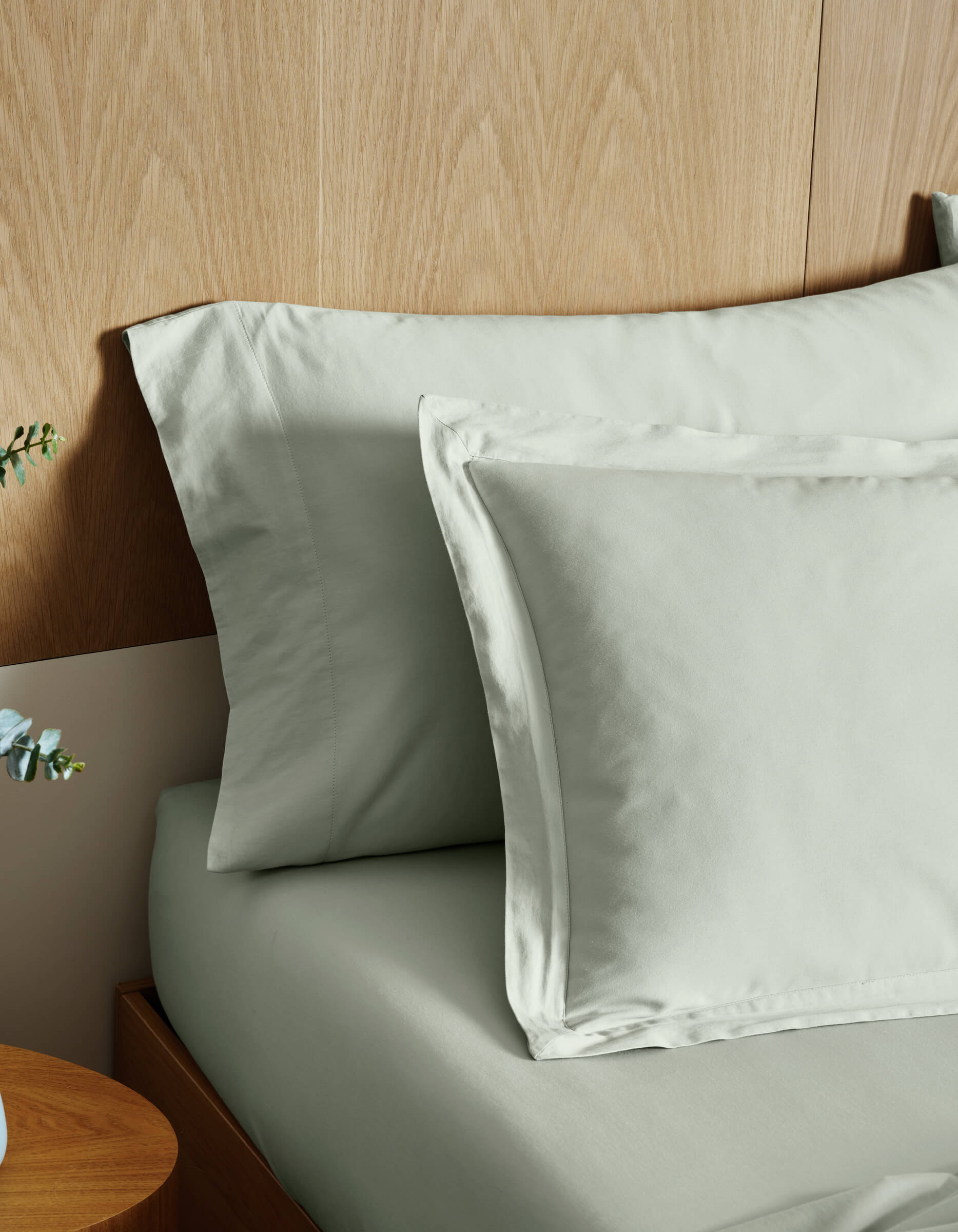Cozy Earth Soft-Wash Cotton Shams in light gray are neatly arranged on a wooden bed with a wood-paneled headboard; part of a wooden nightstand and a small plant appear in the foreground.