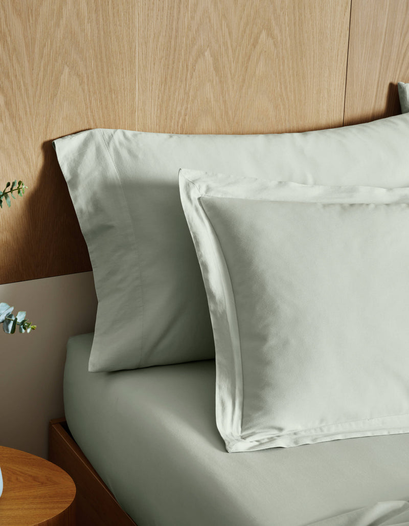 Cozy Earth Soft-Wash Cotton Shams in light gray are neatly arranged on a wooden bed with a wood-paneled headboard; part of a wooden nightstand and a small plant appear in the foreground.