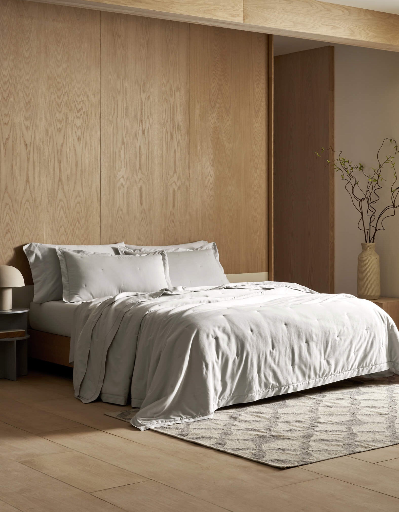 A modern bedroom features light wood-paneled walls, a neatly made bed with the Cozy Earth Soft-Wash Cotton Quilt in light gray, a bedside table with a lamp, a patterned rug, and a tall vase with branches in the corner.