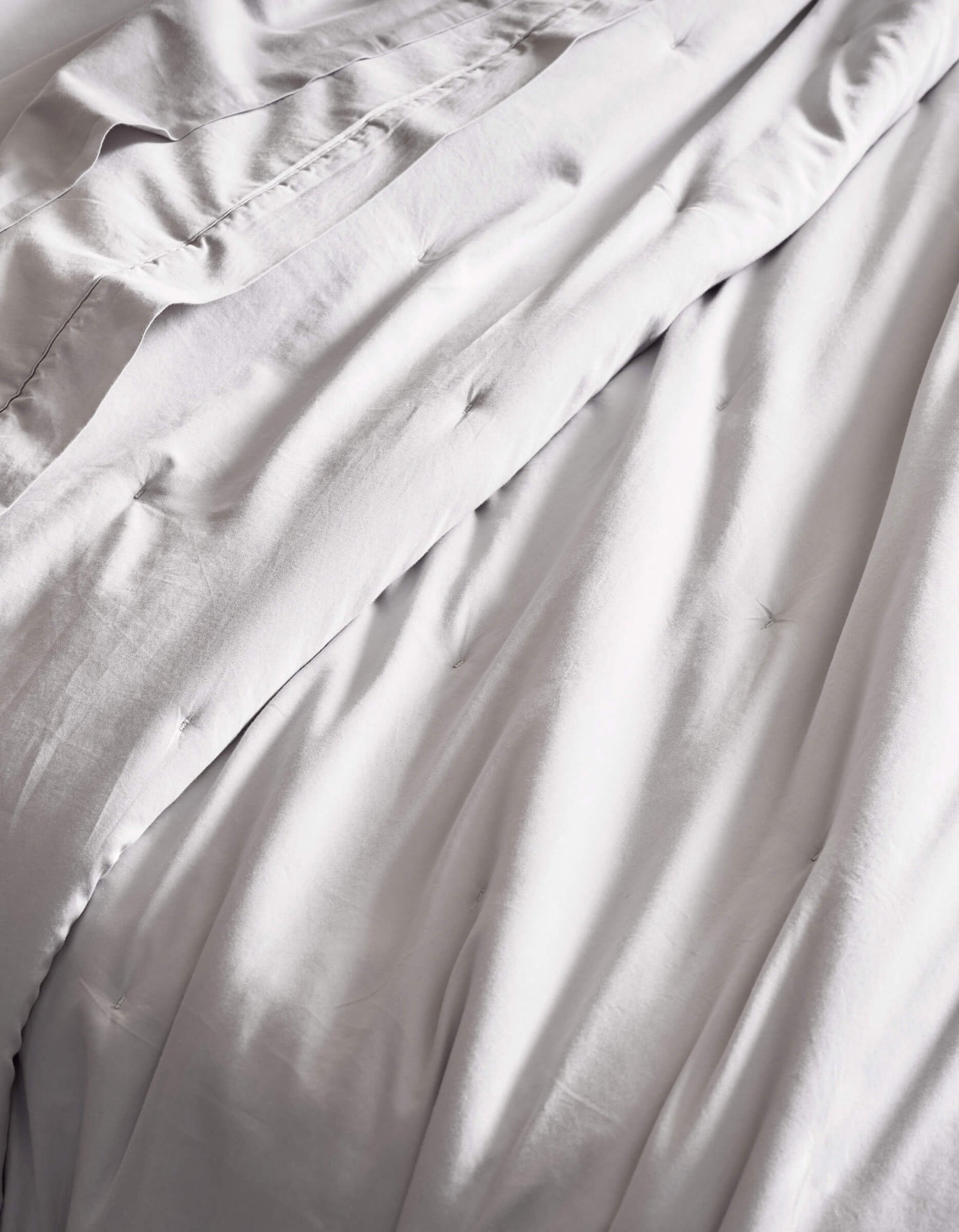 The Cozy Earth Soft-Wash Cotton Quilt features smooth, light gray bedding with gentle folds and subtle tufted details, offering a soft, inviting texture.