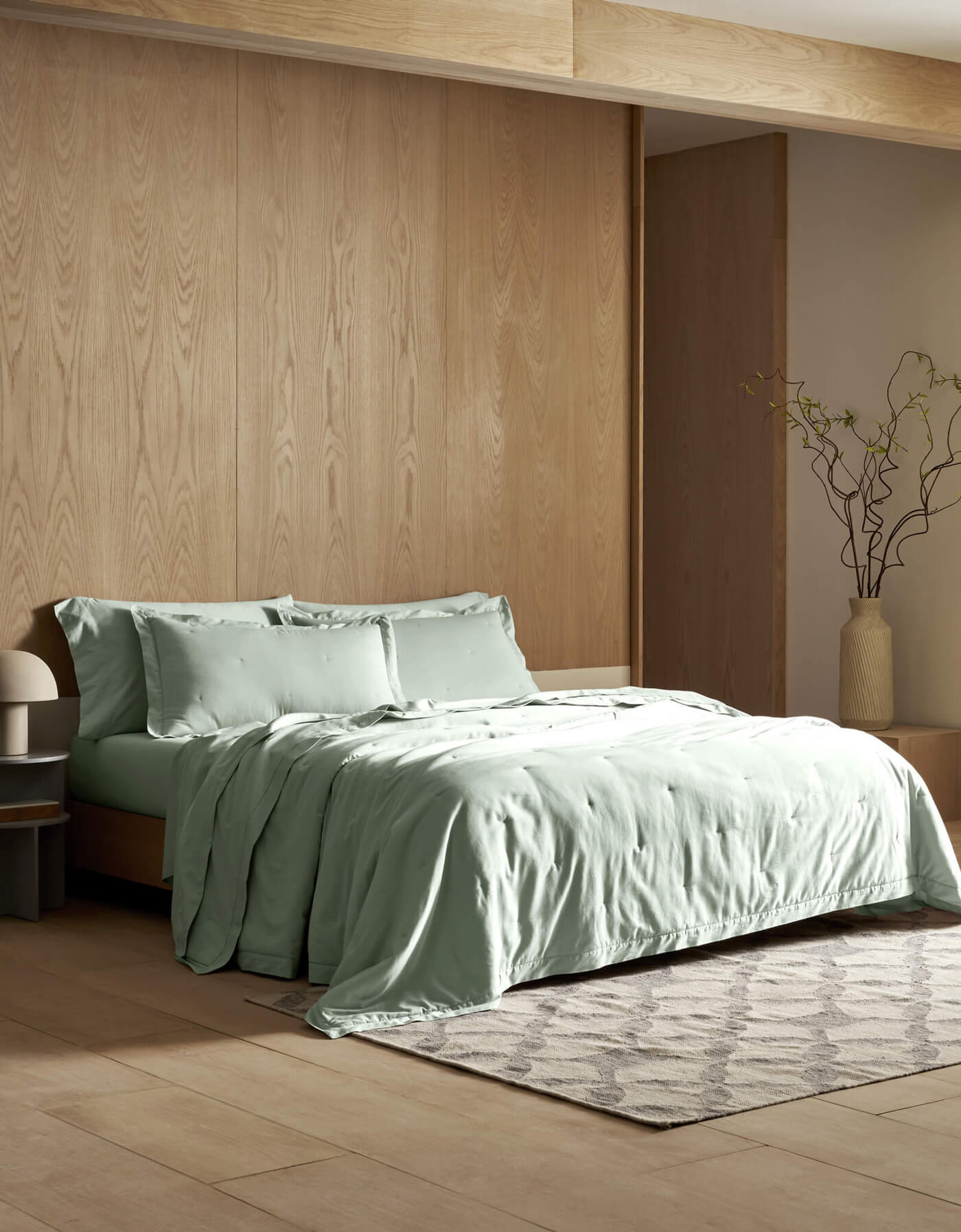 A modern bedroom featuring light wood walls, a bed dressed in Cozy Earth's Soft-Wash Cotton Quilt in sage green, a gray patterned rug, a tall vase with branches, and sunlight filtering through sheer curtains.