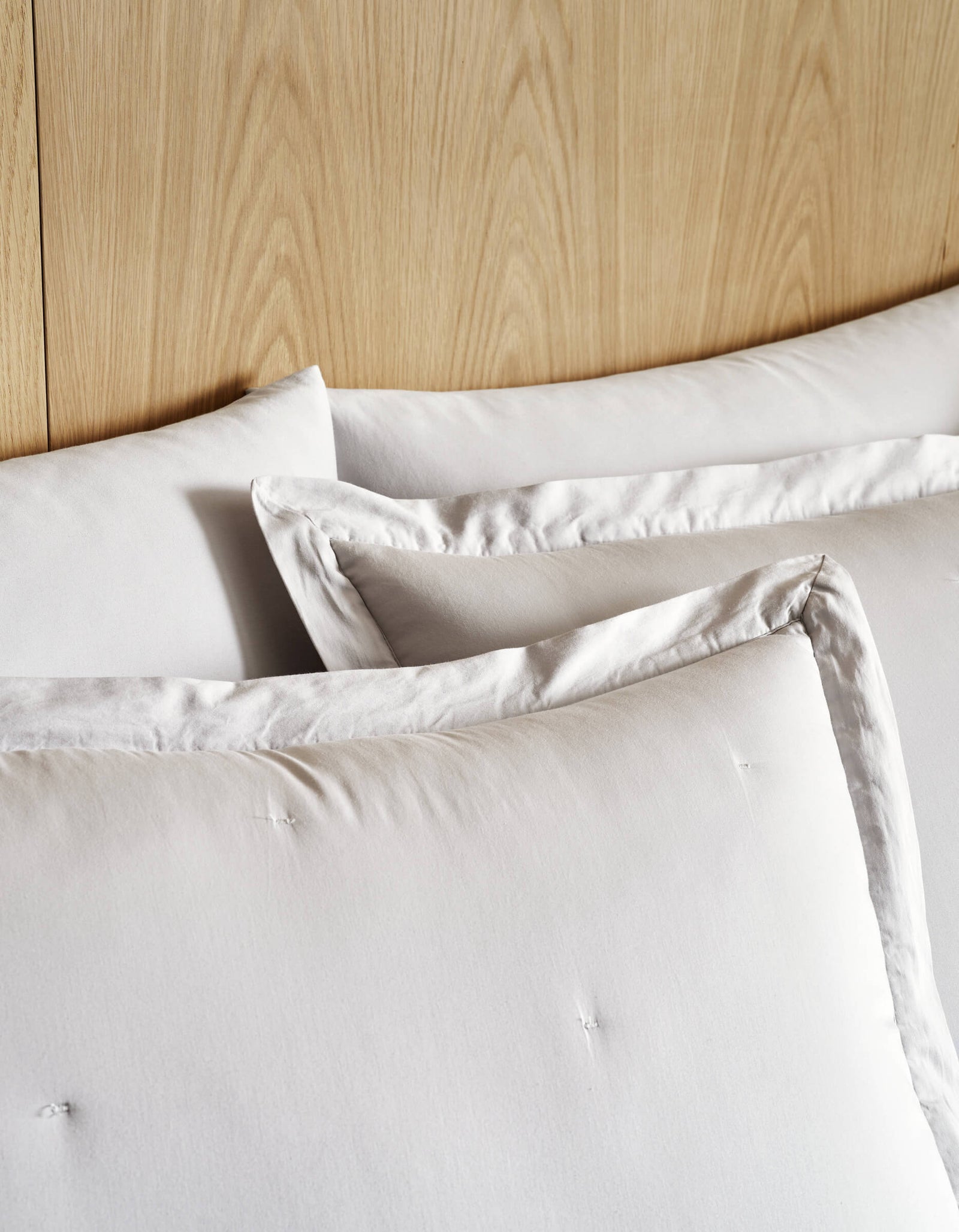 Three large Soft-Wash Cotton Quilted Shams by Cozy Earth, in white with subtle tufting, are stacked against a light wood headboard for a cozy, minimalist bed setting.