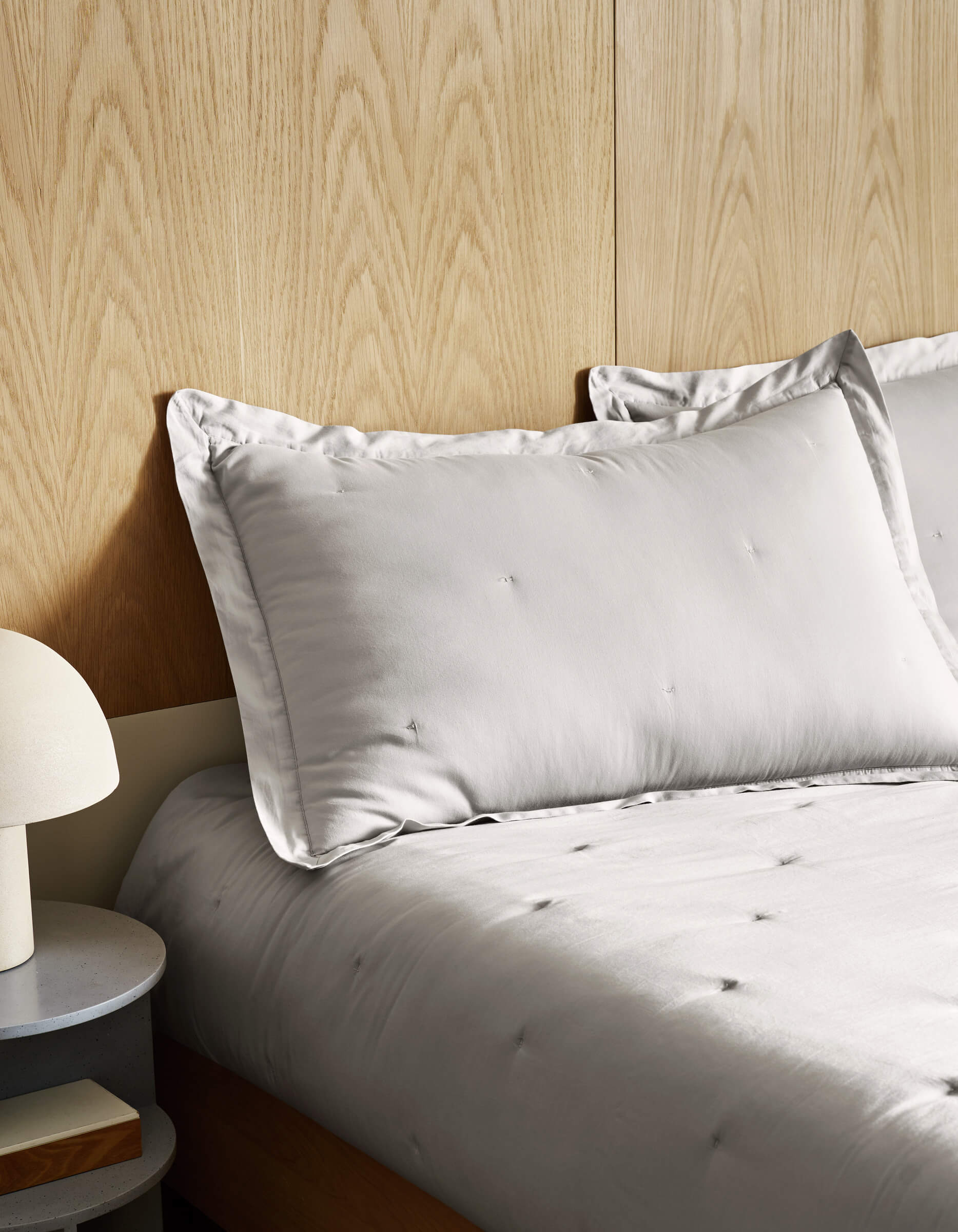 A neatly made bed features the Cozy Earth Soft-Wash Cotton Quilt Set in light gray, with matching pillows against a light wood-paneled wall. On the left, a round bedside table holds books and a white lamp.