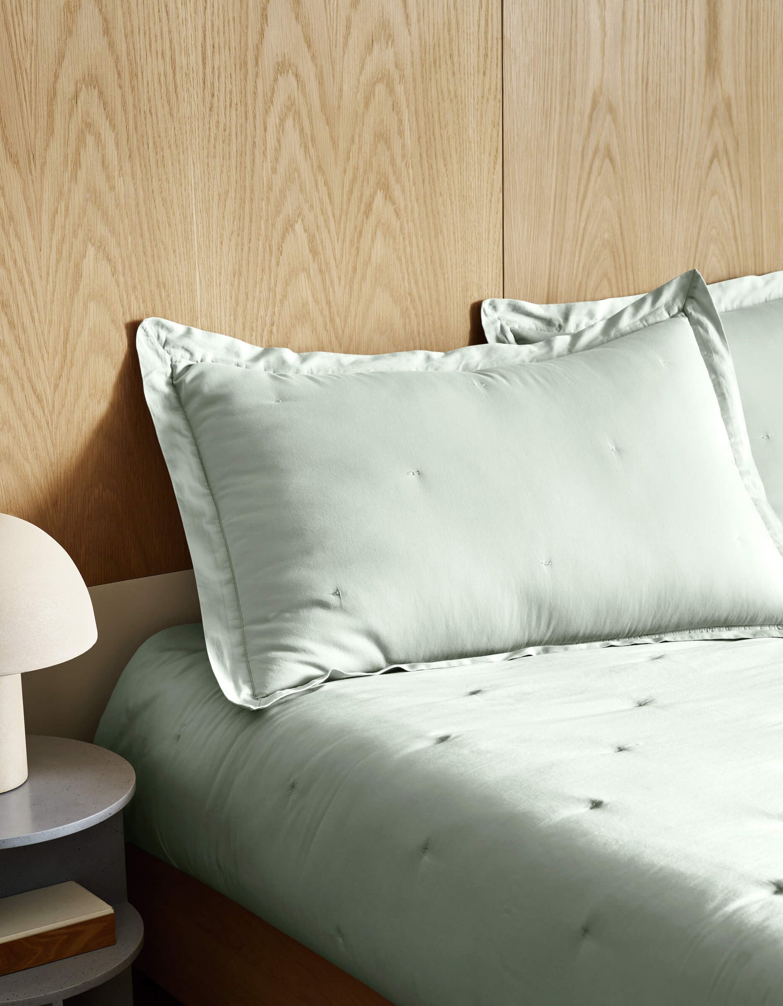 A neatly made bed features Cozy Earth's Soft-Wash Cotton Quilt in light sage green, set against a wooden paneled wall. On the left, a round bedside table holds a modern white lamp.