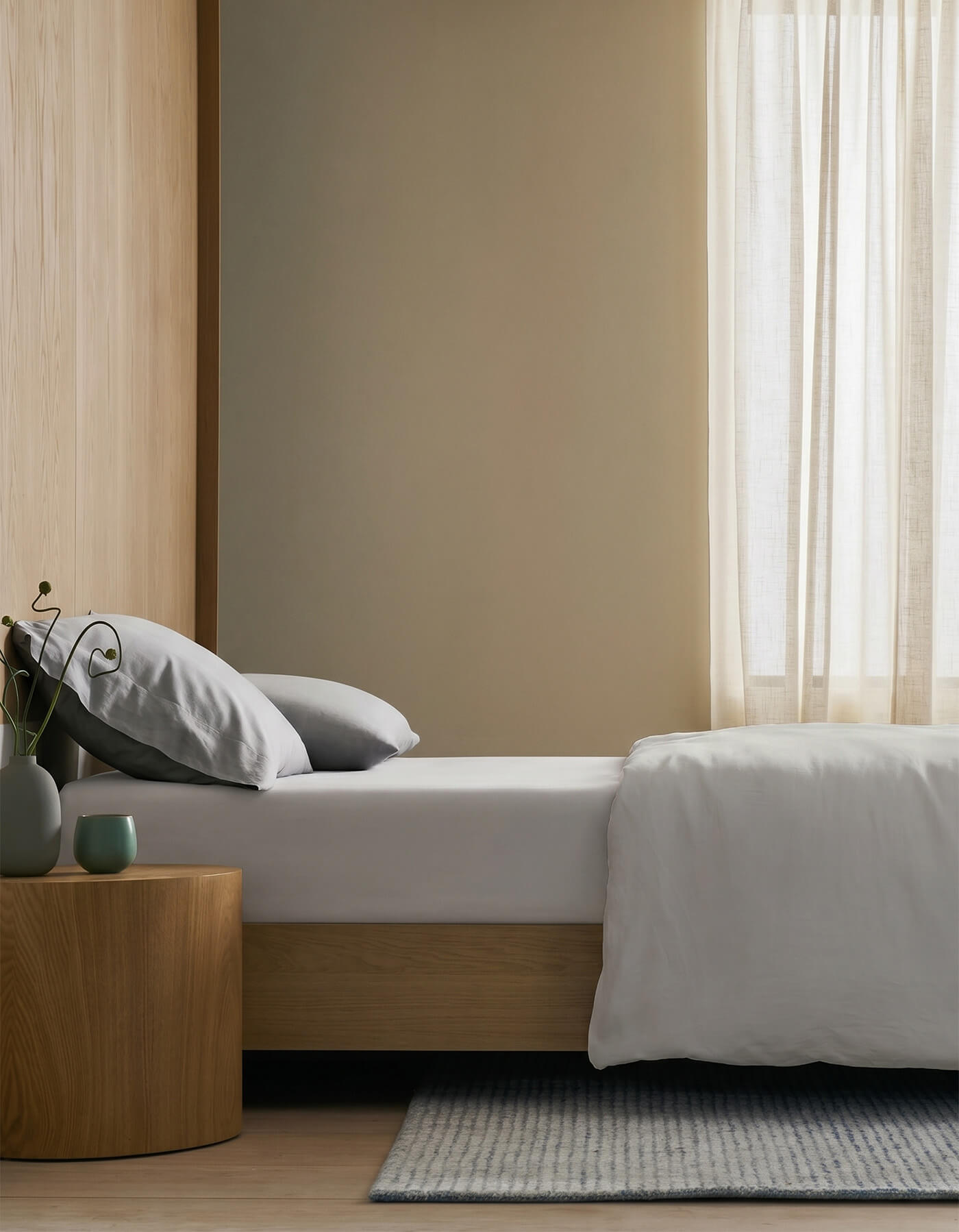 A minimalist bedroom features a low wooden bed with Cozy Earth’s Soft-Wash Cotton Fitted Sheet, gray pillows, white bedding, a wooden nightstand with vases and a plant, light curtains, and a striped rug on light wood floors filled with natural light.
