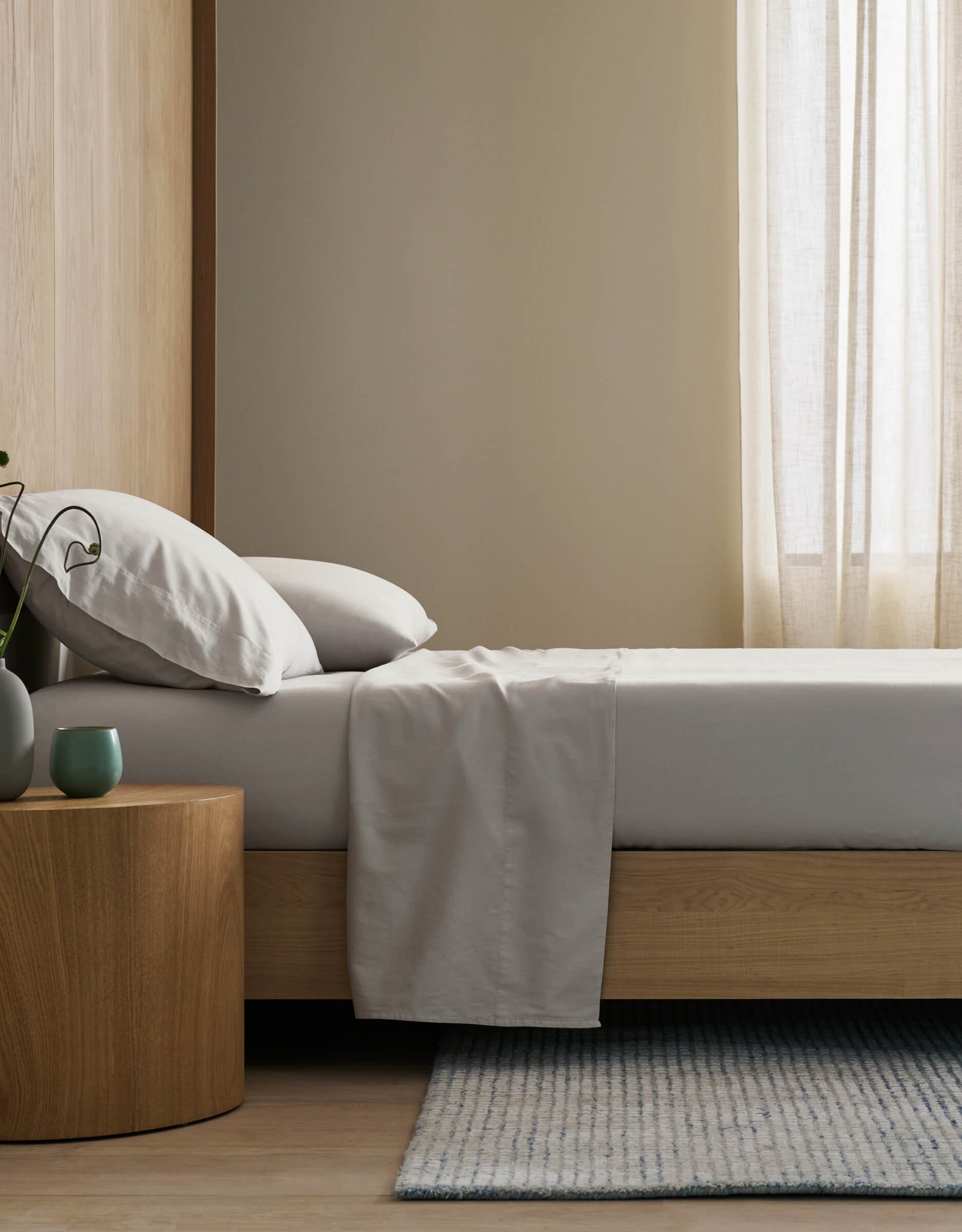 A minimalist bedroom features a neatly made bed with Cozy Earth's Soft-Wash Cotton Flat Sheet, light gray bedding, a wooden nightstand with a green vase and cup, a light blue-gray rug, and sheer curtains letting in gentle natural light.