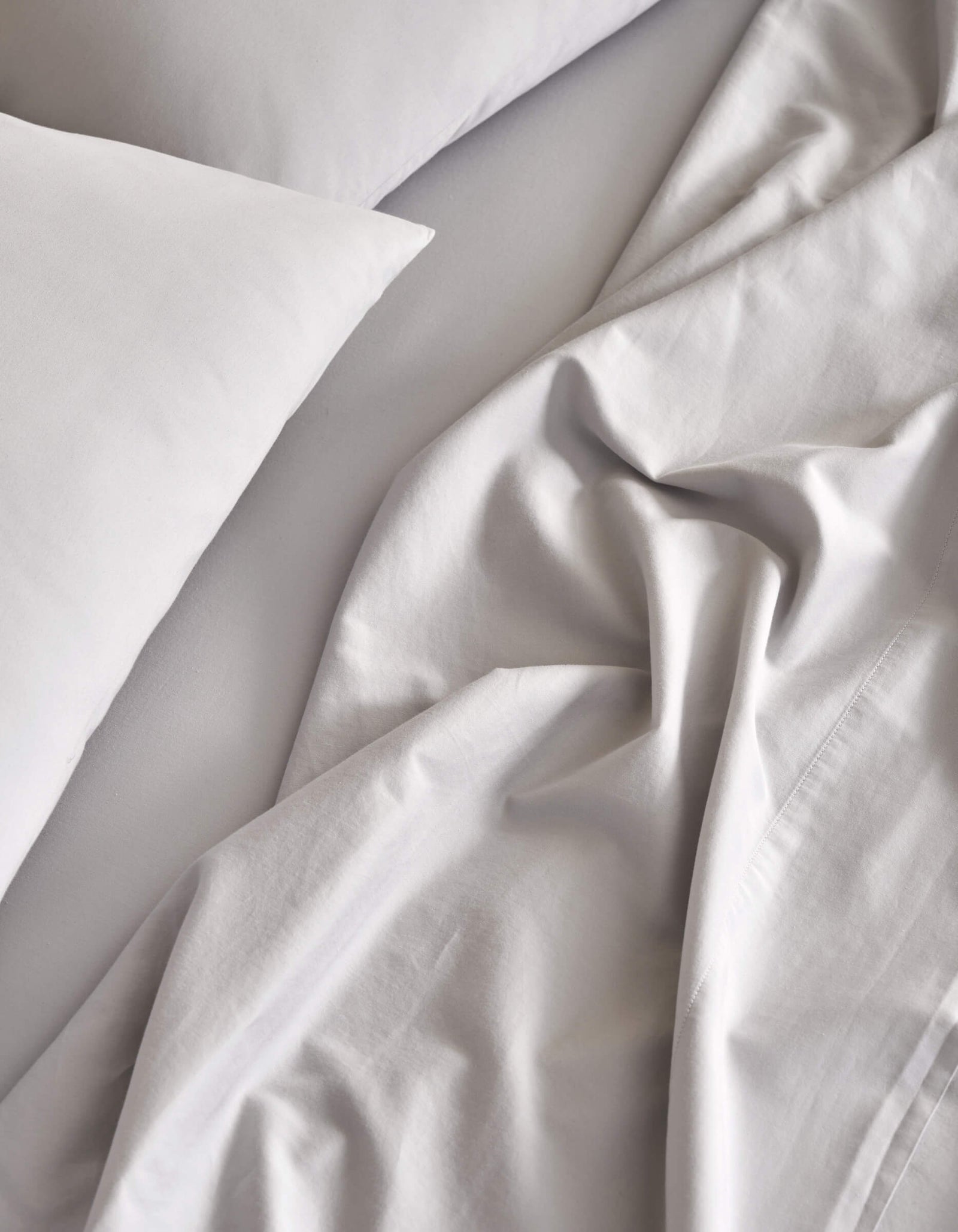 A close-up view of a bed featuring Cozy Earth's Soft-Wash Cotton Sheet Set in light gray. The soft, slightly wrinkled fabric creates a relaxed and inviting look.