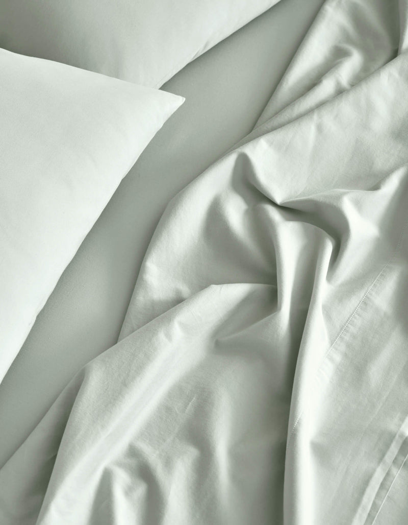 A close-up of a neatly made bed features the Cozy Earth Soft-Wash Cotton Sheet Set in light gray, with slightly rumpled sheets and pillowcases creating soft folds and natural shadows.