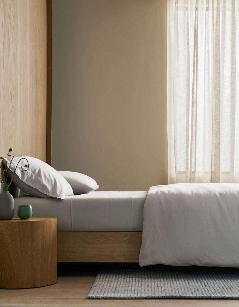 A minimalist bedroom features a neatly made bed with Cozy Earth's Soft-Wash Cotton Bedding Core Bundle, white bedding, and pillows beside a wooden nightstand with a vase and cup. Sheer curtains filter soft natural light, creating a serene ambiance.