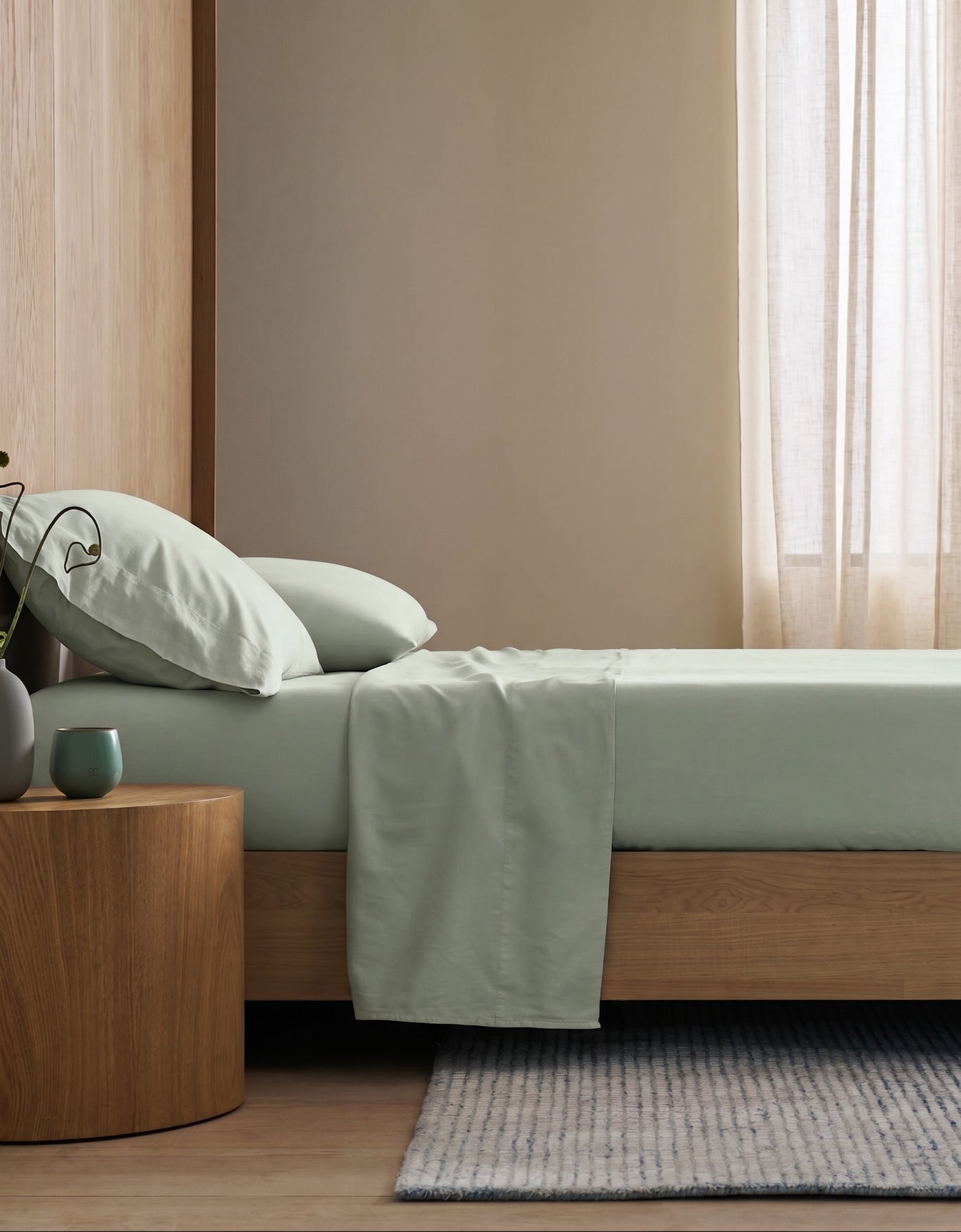 A minimalist bedroom features a wooden bed frame with Cozy Earth's Soft-Wash Cotton Flat Sheet in light green, two pillows, a round wooden nightstand with a green mug and vase, sheer beige curtains, and a light striped rug on wood floors.