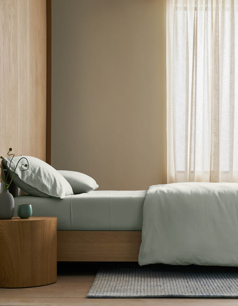 A minimalist bedroom featuring a neatly made bed with Cozy Earth's Soft-Wash Cotton Bedding Core Bundle in pale green, a round wooden nightstand with a plant and cup, light wood floors, and sheer curtains letting in gentle natural light.