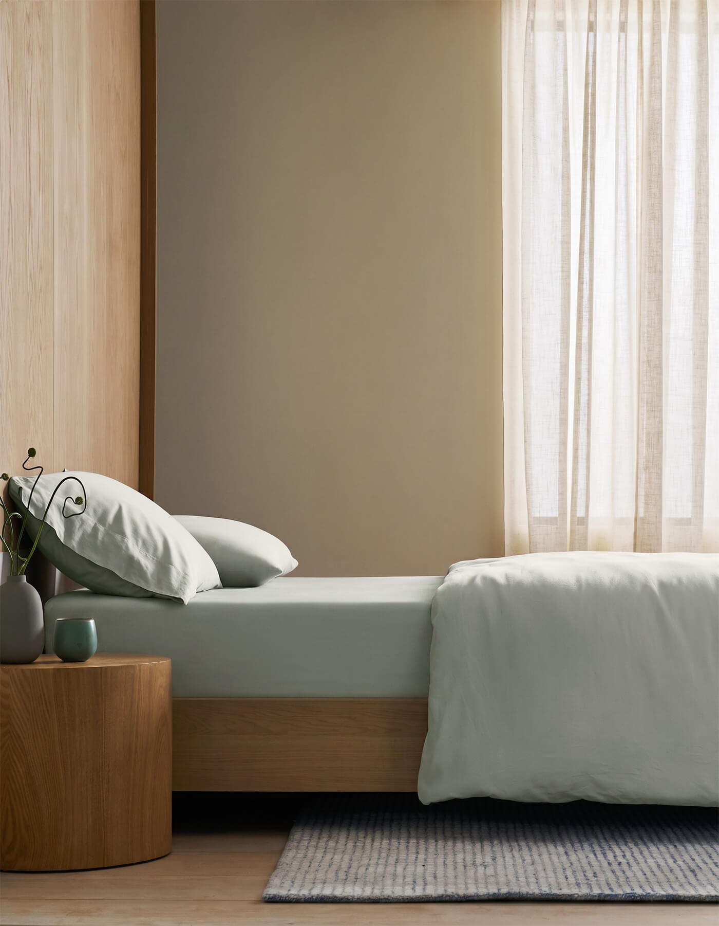 A minimalist bedroom featuring Cozy Earth's Soft-Wash Cotton Fitted Sheet, a light wooden bed, small round wooden side table with a vase, mug and plant, pale gray bedding, light curtains, and a striped rug on the floor.