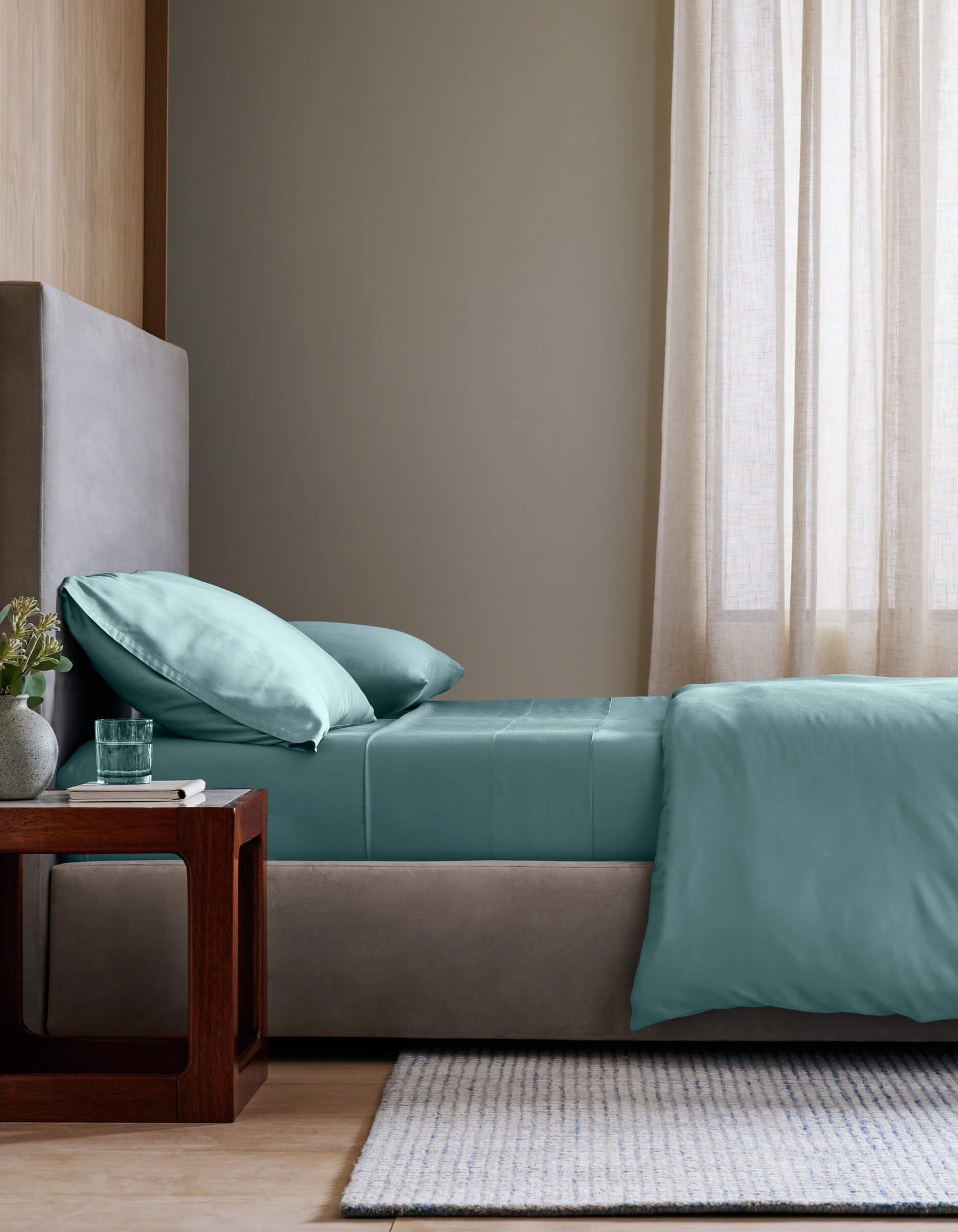A modern bedroom features a neatly made bed with Cozy Earth's Bamboo Sheet Set in teal and two pillows, next to a wooden nightstand holding a glass of water and a small potted plant. Soft natural light filters through sheer beige curtains.