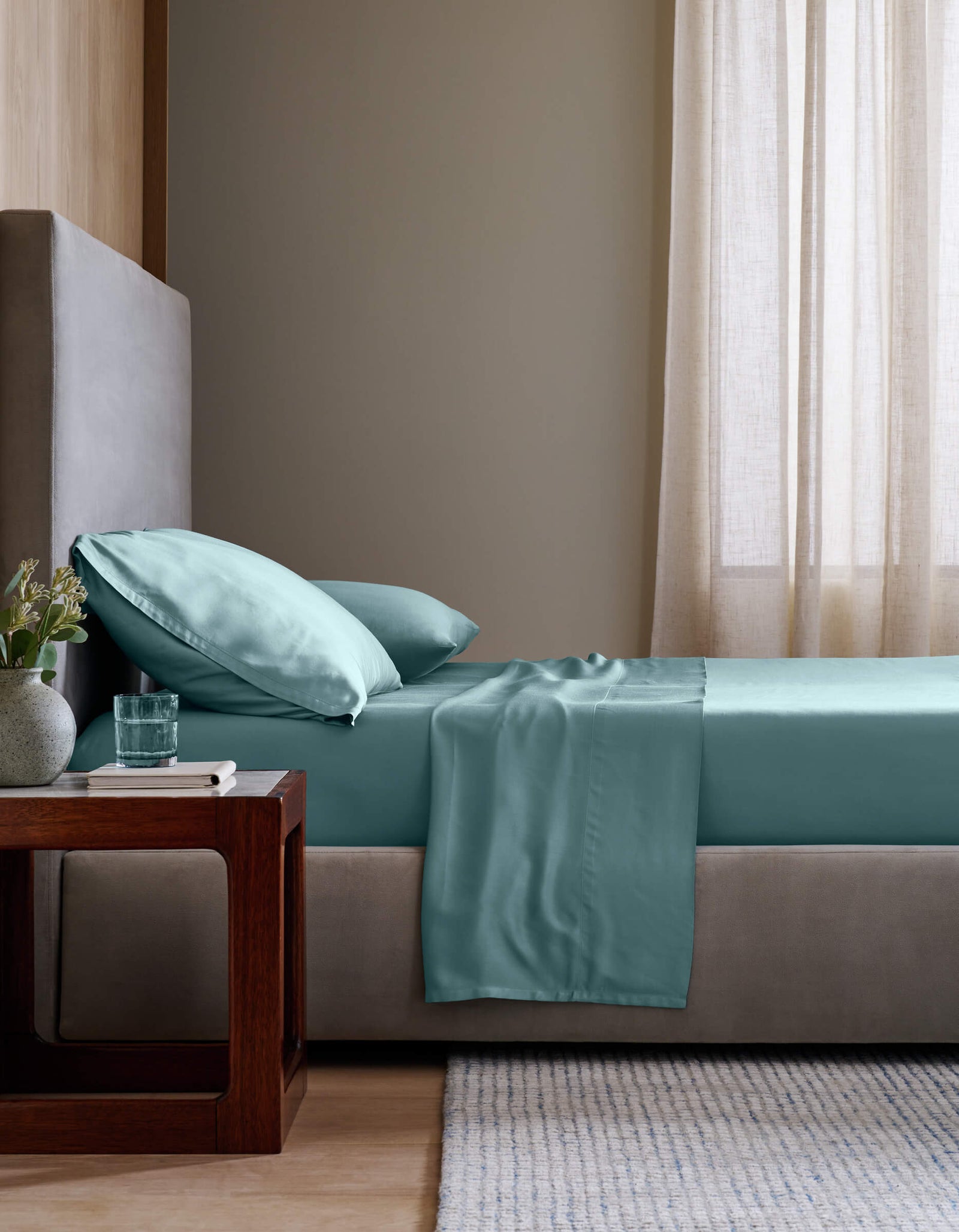 A neatly made bed features Cozy Earth’s Bamboo Flat Sheet in aqua blue, with matching pillows beside a wooden nightstand holding a vase of flowers, a book, and a glass of water. Beige walls and sheer curtains add to the minimalist bedroom style.
