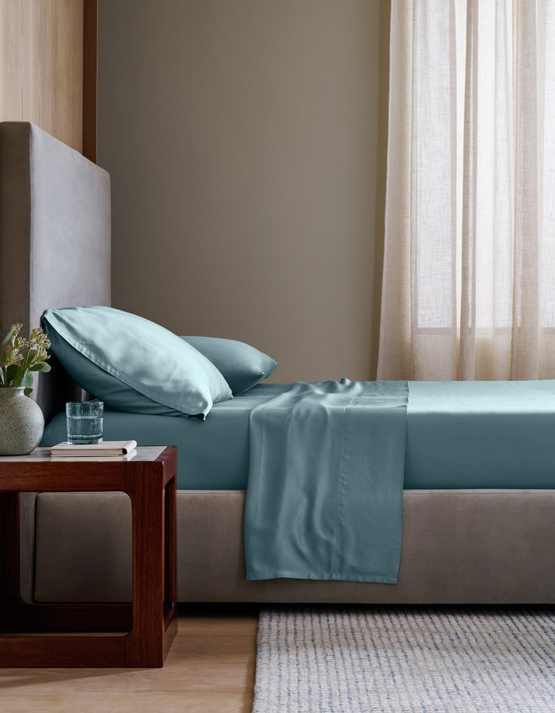 A neatly made bed in a minimalist bedroom setting, featuring a light blue Bamboo Sheet Set set against a neutral gray headboard and beige walls.