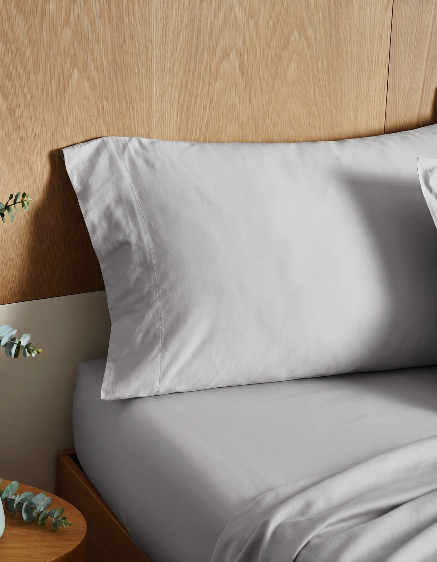 A neatly made bed features Cozy Earth Soft-Wash Cotton Pillowcases in light gray against a wooden headboard, with a small round bedside table and greenery partially visible in the lower left corner.