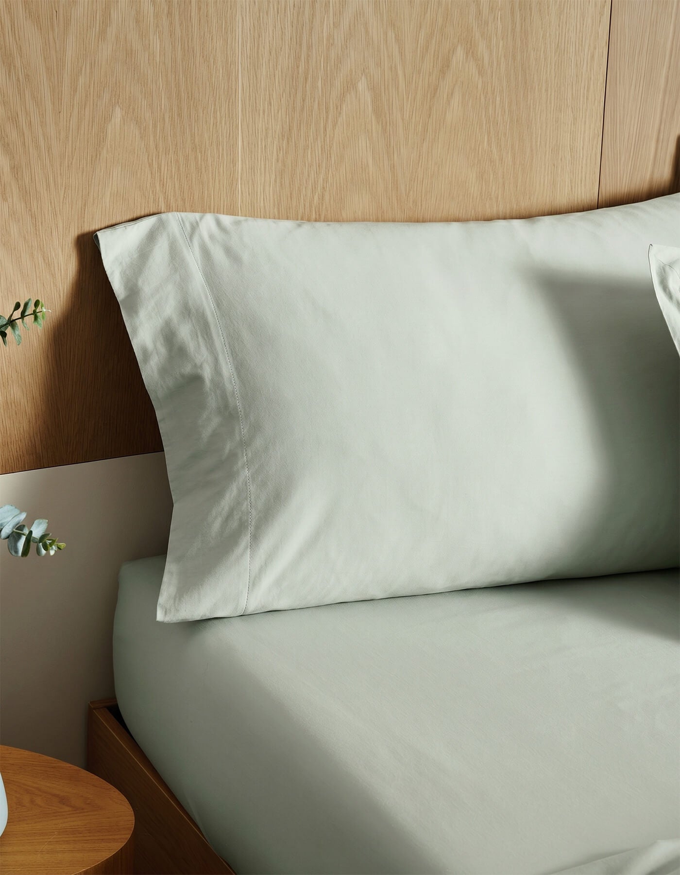 A bed with Cozy Earth's Soft-Wash Cotton Pillowcases in light gray is styled against a light wooden headboard, beside a small wooden nightstand topped with a plant.|Color:Laurel