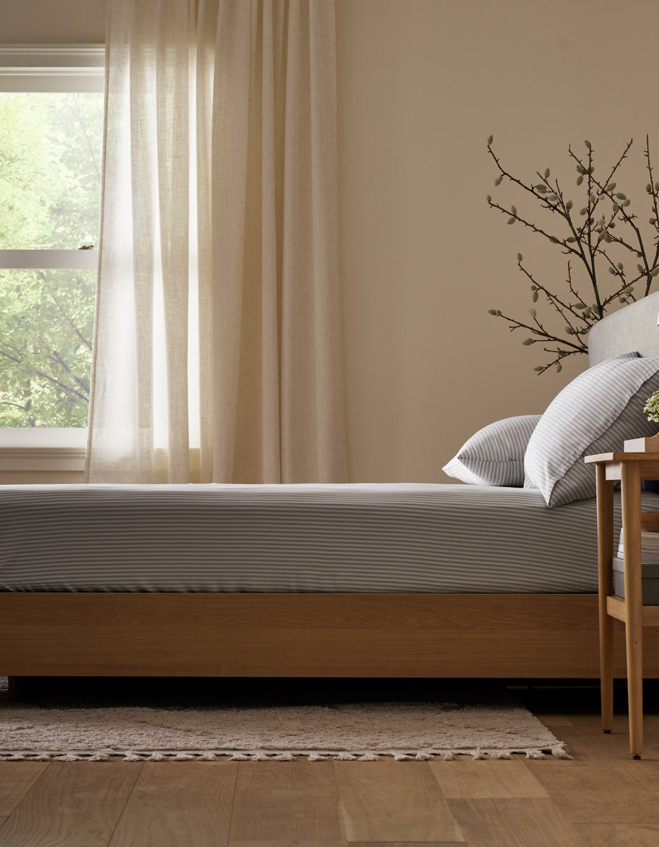 A neatly made bed featuring Cozy Earth’s Bamboo Bedding Basic Bundle sits next to a window with cream curtains, sunlight filtering in to brighten the room. A bedside table and decorative branches add a natural touch.