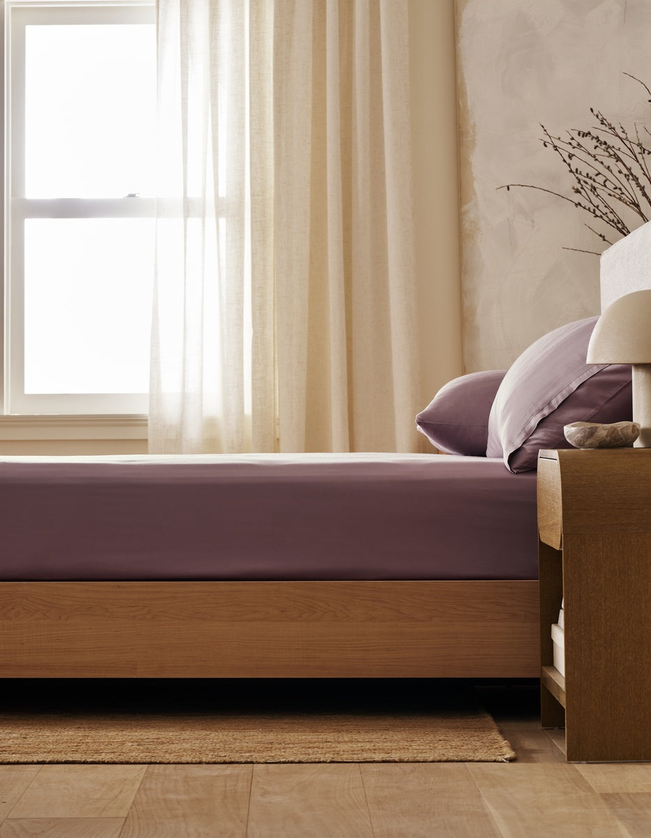 A neatly made bed featuring the Cozy Earth Bamboo Bedding Basic Bundle is positioned beside a wooden nightstand with a small lamp and a vase of branches. Soft light filters through sheer curtains over the window, creating a calm and cozy bedroom atmosphere.
